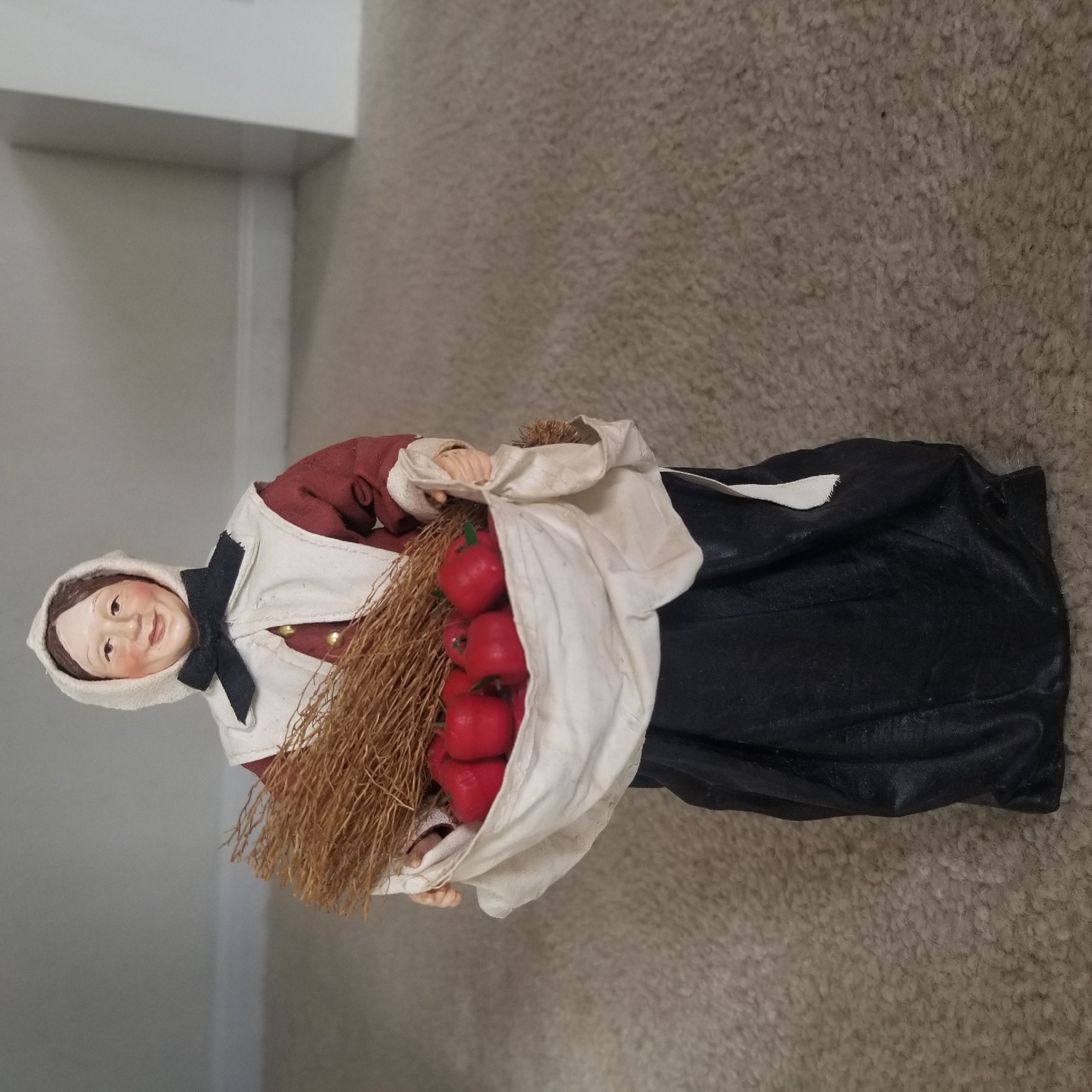 A full shot shows a doll dressed in colonial attire, standing on a carpet. The doll is holding a bundle of straw and a white cloth bag filled with red apples. The doll has a painted face with rosy cheeks and a smiling expression. She wears a white bonnet and apron over a dark red dress and a black skirt. The background is a neutral gray wall with a white baseboard. The doll is positioned centrally in the frame, with the carpet filling the foreground and lower portion of the image. The lighting is even and diffused, suggesting indoor lighting. The location is the city of Virginia Beach, United States of America.