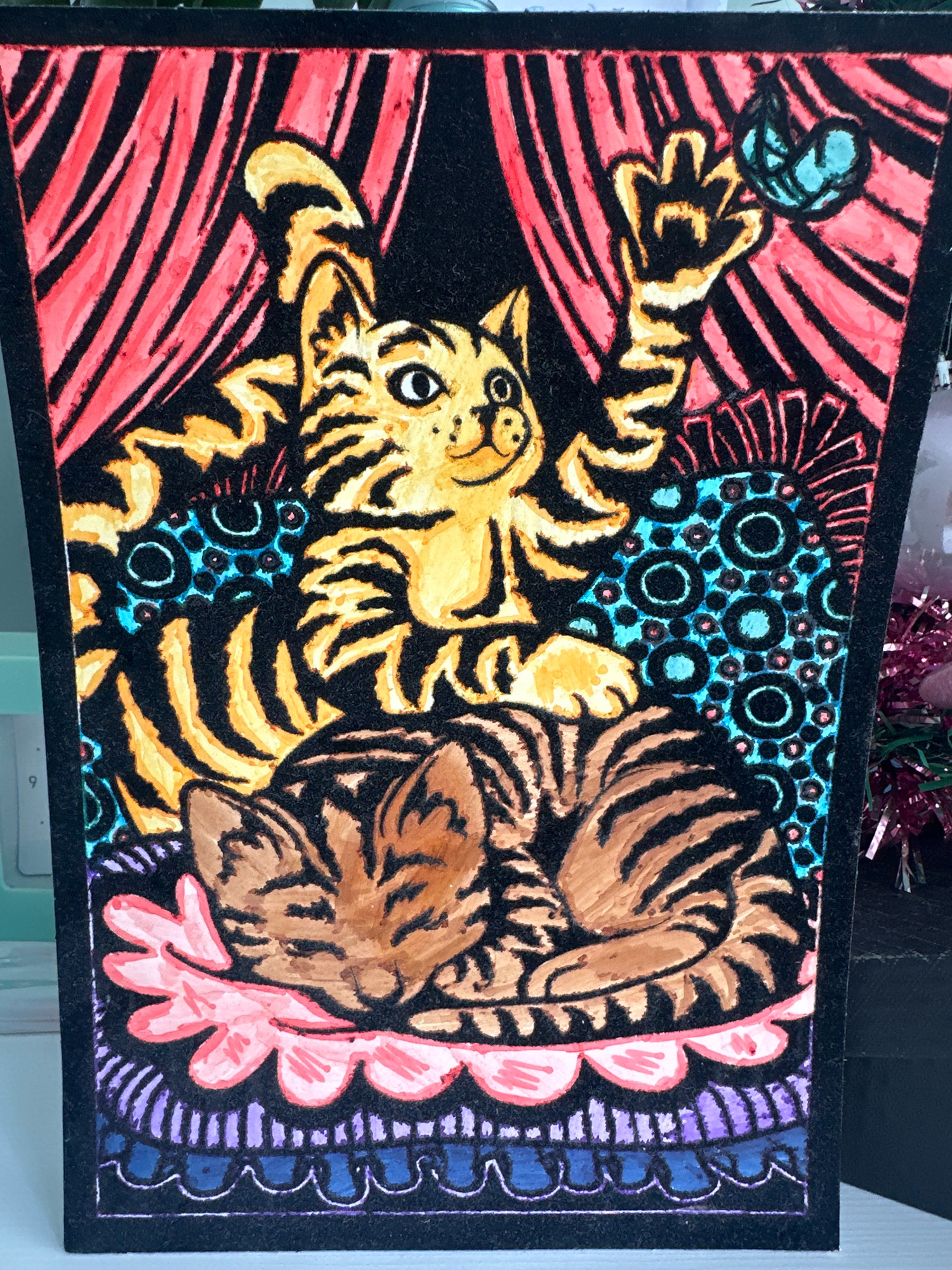 The image is a close-up, eye-level shot of a colorful, decorative piece featuring two stylized tiger-striped cats. The artwork is rendered in a bold, graphic style with vibrant marker colors and black outlines on what appears to be black flocked paper or cardstock.
In the foreground, a brown and black striped cat is curled up asleep on a pink, petal-like cushion. The cushion sits on a decorative border with bands of purple and blue.
Behind the sleeping cat, a yellow and black striped cat is depicted sitting up. This cat is looking towards a small, blue butterfly with outlined wings, which is positioned in the upper right corner of the artwork. The yellow cat has one paw raised, reaching towards the butterfly. The background behind the cats features red and black striped curtains. The cats are positioned on a patterned surface with blue circles outlined in black.
The artwork is framed by a thick black border. The overall impression is that of a playful and decorative art piece, possibly a greeting card or a small decorative panel.
There is no information in the image to suggest its location in the city of Minsk, Belarus.