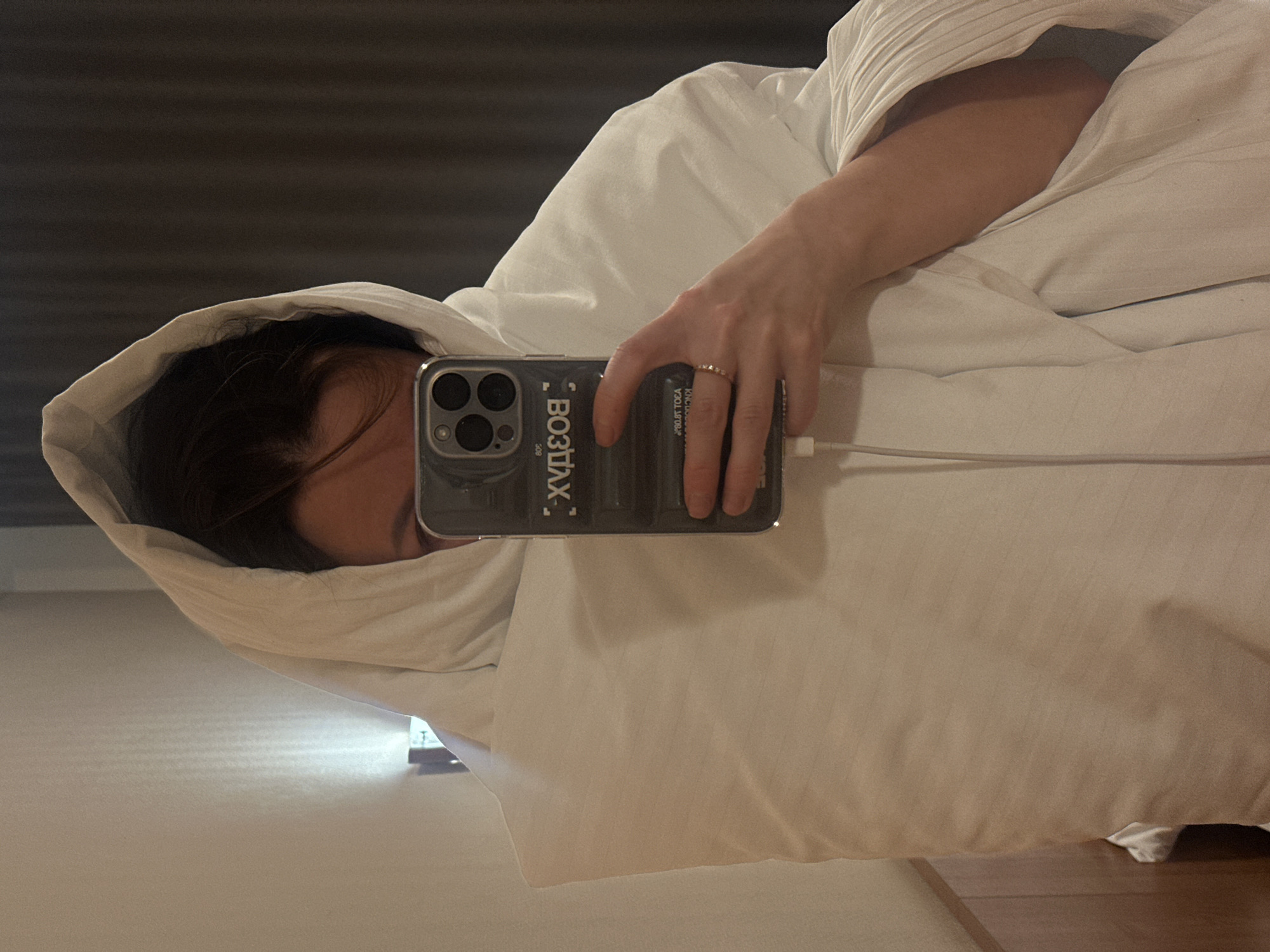 A person is taking a selfie with an iPhone while wrapped in a white blanket. The phone has a clear case with Russian text "ВОЗДУХ" on it. The person is wearing a ring on their finger. The background appears to be a bedroom interior. There is no information available to confirm the location of this image to be Perm, Russia.