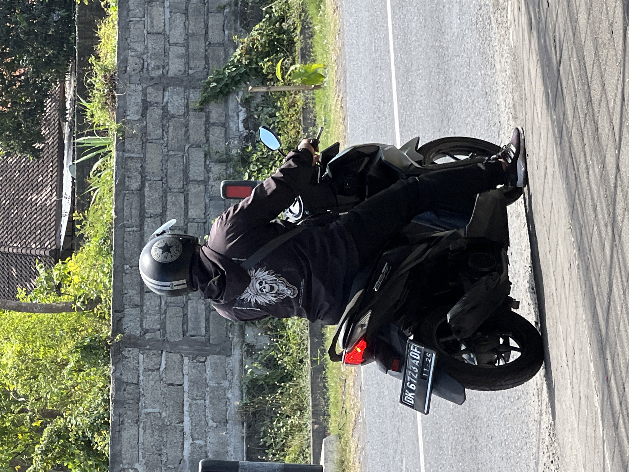 A person wearing a black hoodie with a skull graphic and a black helmet with white stripes is riding a black scooter. The scooter has a license plate that reads "DK 6723 ADF" and "11-25". The scooter is on a paved road next to a sidewalk. In the background, there is a low brick wall, vegetation, and a tiled roof of a building. The scene appears to be in Denpasar, Indonesia, given the license plate format.
