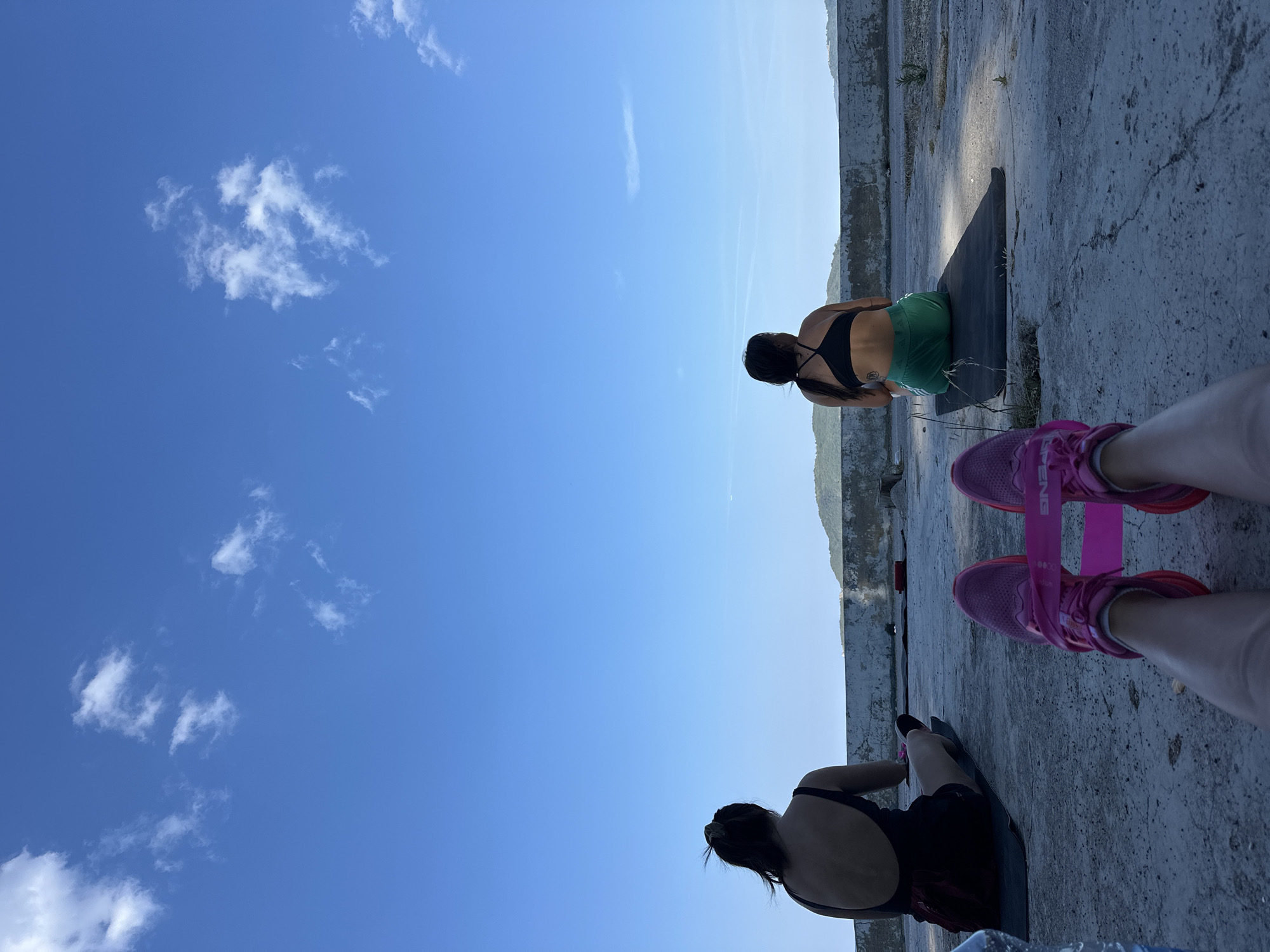 Two individuals are seated on yoga mats outdoors, facing away from the camera, with their backs to a serene blue sky dotted with sparse white clouds. The person on the left, wearing a black athletic top and dark shorts, is positioned on a dark mat. The person on the right, dressed in a black sports bra and green shorts, sits on another dark mat. In the foreground, the legs of a third person are visible, wearing bright pink sneakers with a pink resistance band stretched between them. The ground is a textured concrete surface with some scattered vegetation. In the background, a low concrete wall separates the foreground from a landscape of rolling, green hills that recede into the distance under the expansive sky. The scene suggests an outdoor workout or meditation session taking place on a rooftop or elevated area overlooking the bay of Boreti, Montenegro.