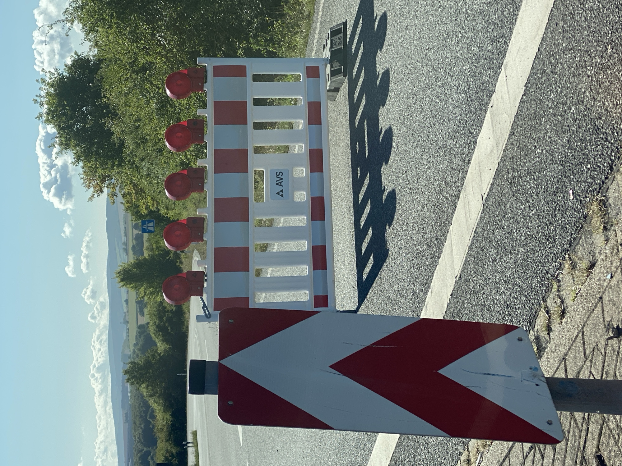 The image shows a temporary road barrier set up on a paved road in Werther, Germany. The barrier is white and red striped, with flashing red lights on top. A chevron sign, also red and white, is positioned to the left of the barrier, indicating a directional change or hazard. The road appears to be a two-lane road, with a white lane divider visible. In the background, there are trees and rolling hills under a partly cloudy blue sky. The scene suggests a road closure or construction area.