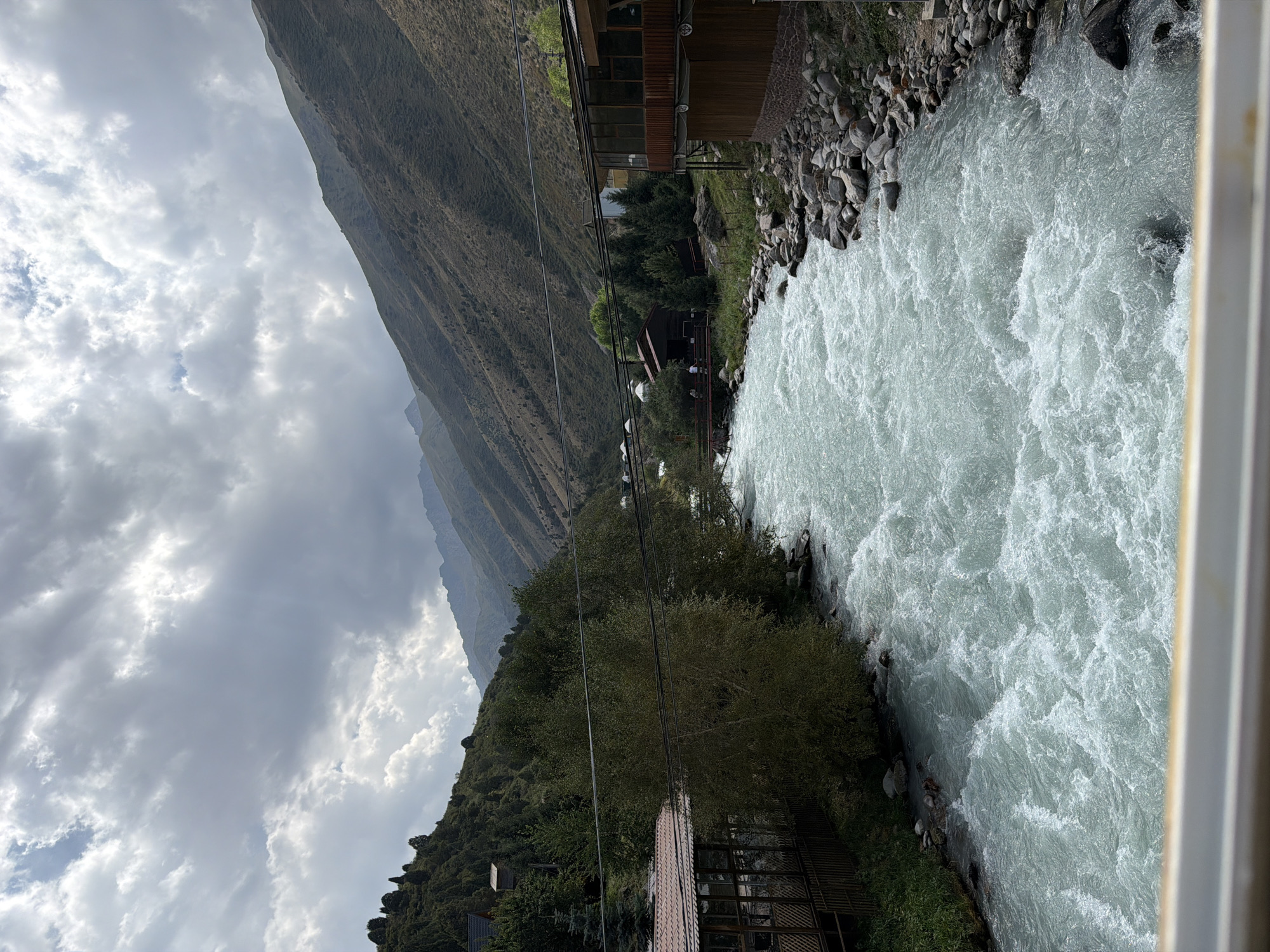 A wide, turbulent river flows through a mountain valley under a cloudy sky. The river, with milky-blue, whitewater rapids, occupies the majority of the frame, moving from the upper left towards the lower right. Along the left bank, a wooded hillside is dotted with some buildings, including a structure with a red roof and an outdoor dining area. The right bank features a rocky shoreline and a building with large windows and a wooden balcony. In the background, steep, green mountains rise towards the horizon, their slopes covered in sparse vegetation. Several white yurts are visible in the distance on the left side of the valley. Overhead, dramatic clouds with visible sunbeams suggest a dynamic weather system. There are no people clearly visible in the image.