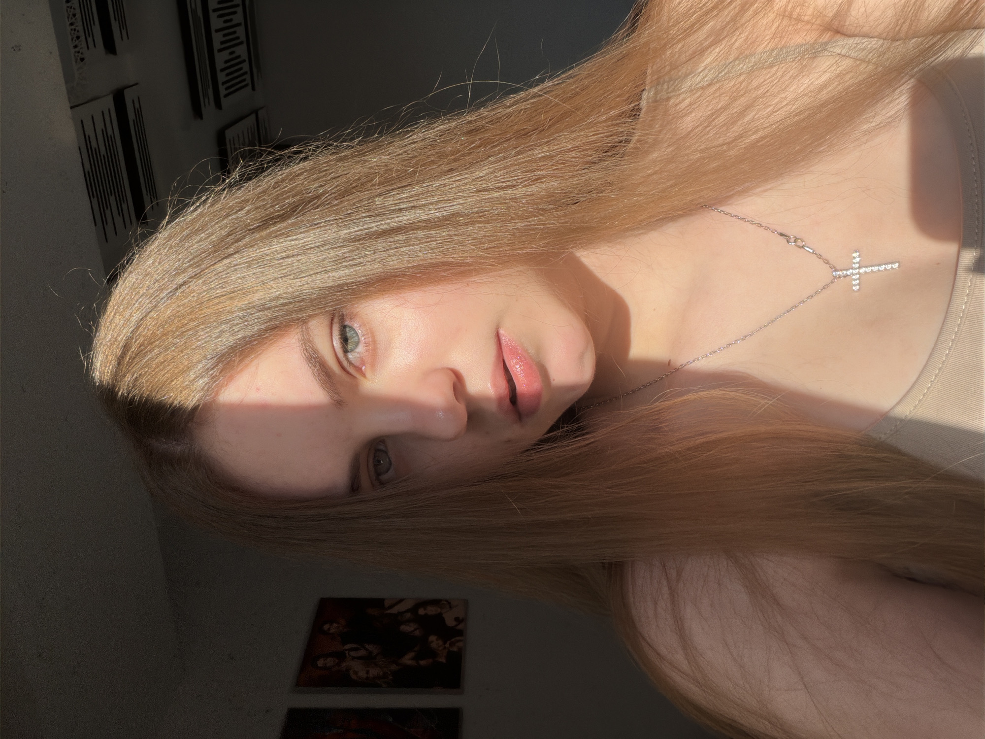 The image shows a young Caucasian woman with long, straight, light brown hair. She is looking directly at the camera with light blue eyes and wearing a beige top and a silver cross necklace. A shadow is cast diagonally across her face, obscuring the left side of her forehead and eye. The background is dimly lit, with two framed pictures visible on the left wall and acoustic foam panels on the ceiling. The scene appears to be indoors, possibly in a residential setting in Dnipro, Ukraine, given the implied context of the request. The lighting suggests it is daytime, with sunlight entering from the right.