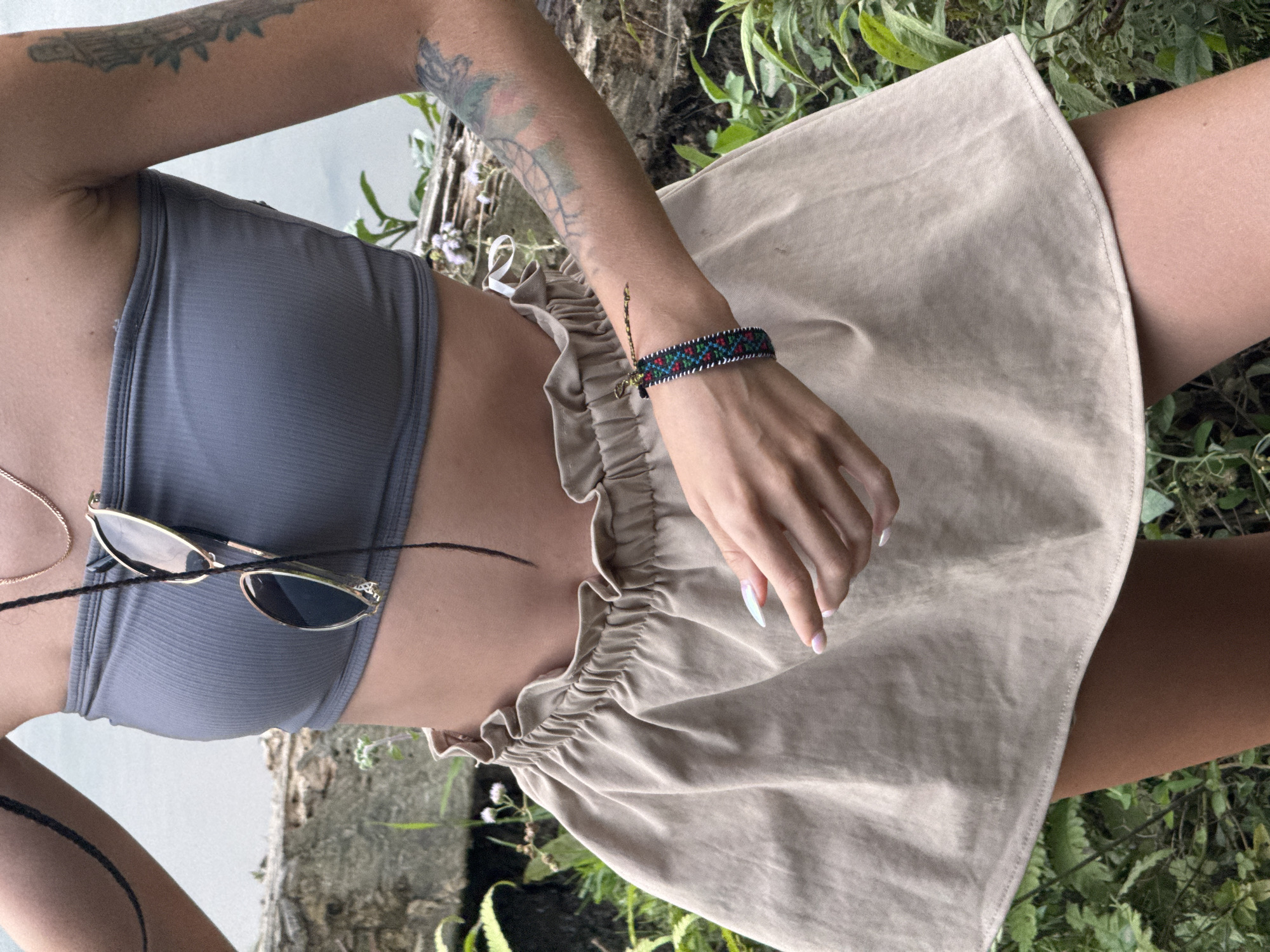 A close-up, eye-level shot, rotated clockwise, captures a person from the mid-torso to the upper thighs, set outdoors in what appears to be a natural, green environment. The person wears a simple grey bandeau or crop top and a light beige or khaki skirt with a ruffled, elastic waistband. Gold-framed sunglasses are casually tucked into the top. A delicate gold chain necklace is visible on the left.
The person's right arm is prominent, adorned with multiple tattoos, including leafy patterns and a design resembling a dreamcatcher. A colorful woven or beaded bracelet with red, blue, and black geometric patterns is worn on the right wrist. The hand, with long, pointed, light-colored manicured nails, rests gently on the skirt.
The background is dominated by lush green foliage and plant life, indicative of a natural landscape. A textured tree trunk or wooden structure is visible on the right, while a lighter, more distant surface, possibly water or a wall, is seen on the left. The bright, even lighting suggests it is daytime. The overall scene conveys a relaxed, summery feel, consistent with an outdoor setting in a location like Nậm Dịch Commune, Vietnam. No text is visible in the image.