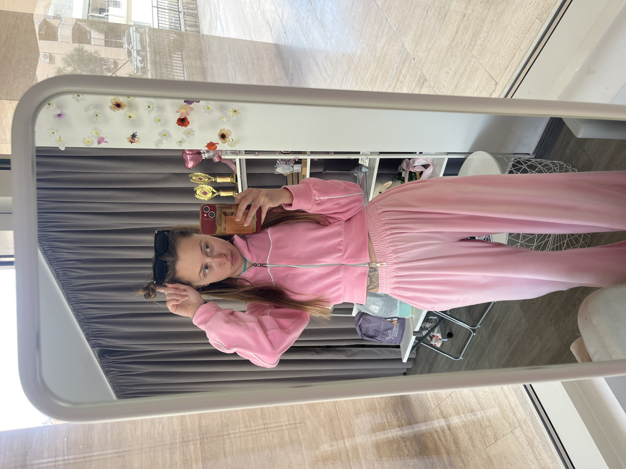 A fair-skinned woman is reflected in a rectangular, white-framed mirror, taking a selfie with a cell phone in her right hand. She is wearing a pink tracksuit with white trim. Her light brown hair is partially pulled back, and she is wearing sunglasses on her head.
Behind her in the reflection are gray curtains, a white bookshelf unit with various items including floral decorations, and other furniture including a white chair with a metal frame. The floor is light-colored tile.
Outside the mirror's frame, tiled flooring extends to a window. The reflection in the window suggests an exterior urban scene, possibly in Budva, Montenegro, based on building styles.