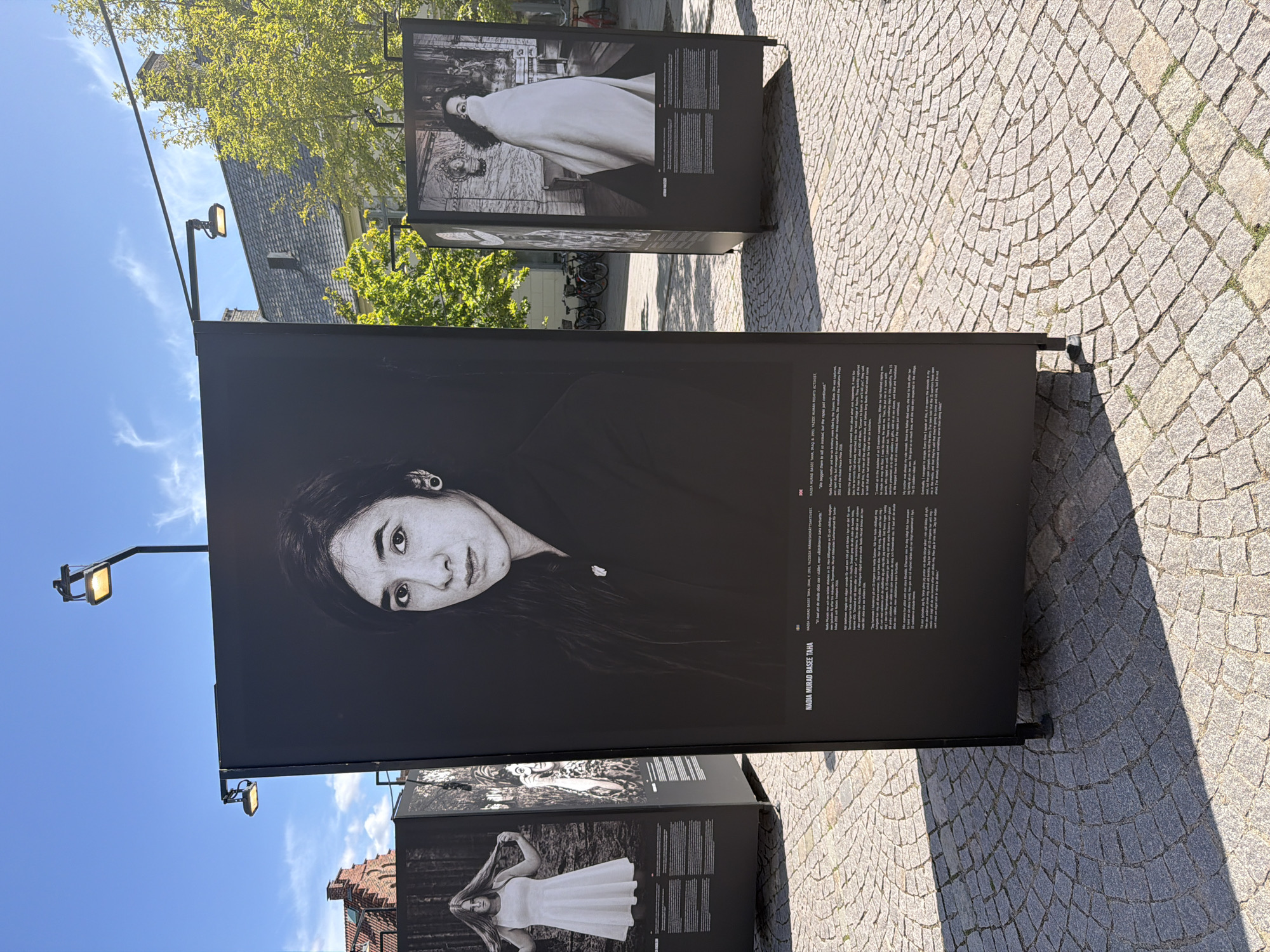 The image depicts an outdoor exhibition of large-format black and white photographs displayed on black panels. The exhibition is set in a paved public space, possibly a square or plaza, in Lund, Sweden. The ground is paved with cobblestones arranged in a concentric pattern. In the background, there are buildings with red tile roofs, trees with green foliage, and a bright blue sky with wispy clouds. Streetlights are visible, mounted on poles extending over the exhibition panels.
The central panel features a portrait of a woman with dark hair and expressive eyes, looking directly at the camera. Text is printed below the portrait, likely providing biographical information and context for the photograph. To the left and right of the central panel, other exhibition panels are visible. The panel on the left shows a woman with long hair, wearing a white dress. The panel on the right shows a woman draped in a white cloth. The overall impression is one of an art installation in a public urban setting.