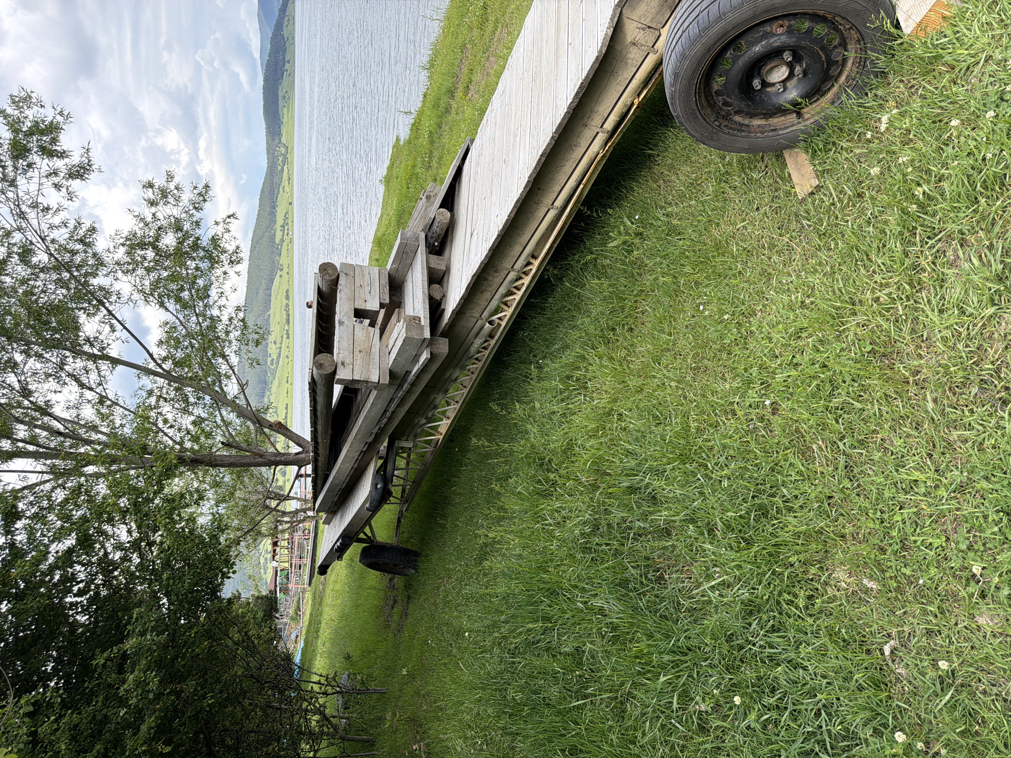 This image, captured in Tsigov chark, Bulgaria, presents an outdoor scene featuring a large, inclined wooden structure positioned on a grassy bank next to a lake. The wooden structure, likely a disassembled pier or boat ramp, is elevated on a metal frame and supported by two visible black tires with rusty rims; one tire is prominent in the bottom right, resting on a wooden block, while another is partially obscured in the mid-left. Various wooden planks and logs are stacked unevenly on top of the main structure. A single individual with dark hair and a dark top is partially visible behind these stacked timbers, looking towards the left side of the lake.
The foreground consists of lush green grass with scattered small white flowers. To the left, dense green foliage from trees and bushes obscures the background. A large tree with a prominent trunk and sparse green leaves stands in the mid-ground, slightly right of center. The smooth surface of a lake occupies the middle and right sections of the image. Across the lake, rolling green hills, partially covered with trees, extend into the distance under a cloudy sky with varying shades of grey and white. In the far distance on the left side of the lake, faintly visible through the trees, are several structures with colorful roofs and a small pier.