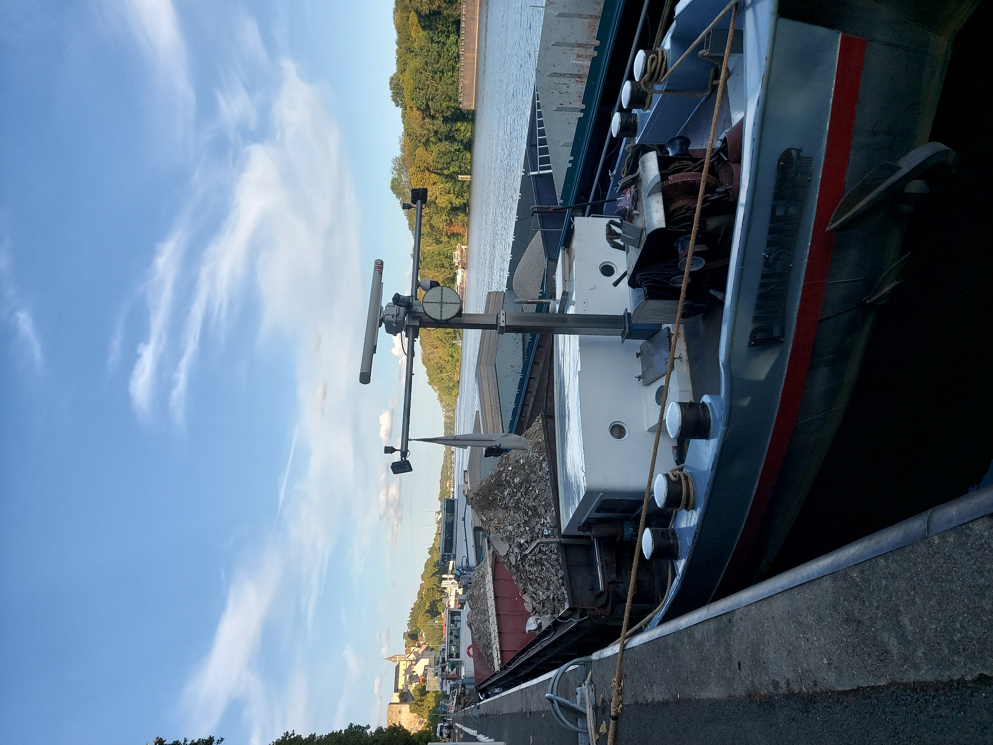 The image depicts a view of the Seine River in Conflans-Sainte-Honorine, France, looking east. In the foreground, a barge named "PUNISHER" is docked along a concrete embankment. The barge is loaded with what appears to be construction debris. Several other barges are moored alongside it. Beyond the barges, the river stretches into the distance, bordered by lush green trees on the right bank. In the background, on the left bank, buildings and a church tower are visible, suggesting the presence of the town. The sky is blue with scattered white clouds.