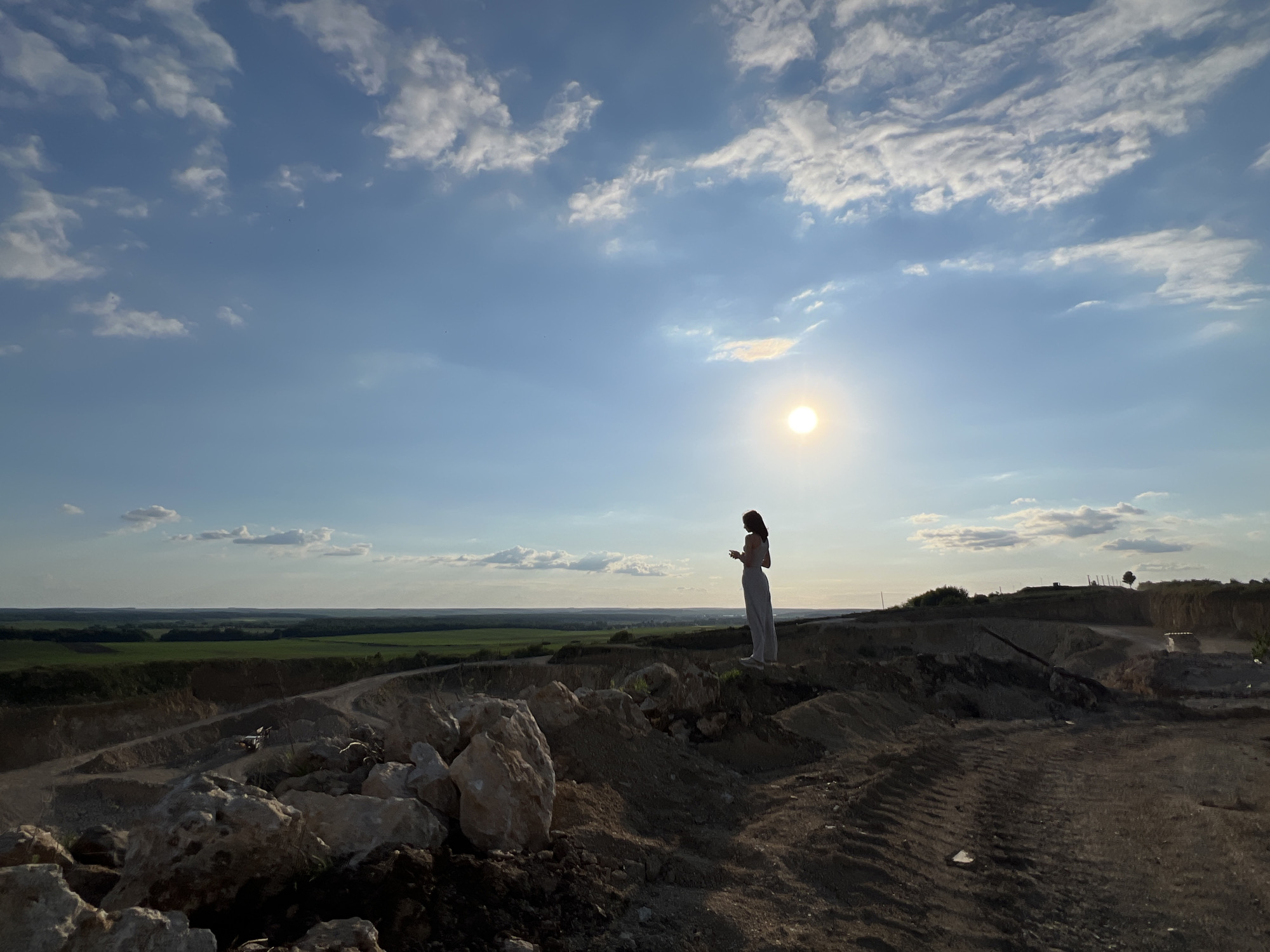 A solitary woman stands on a mound of earth, silhouetted against a bright sun in a hazy sky. She is wearing a long-sleeved top and loose pants. The foreground is dominated by piles of rocks and dirt, with tire tracks visible on the ground. In the background, a vast landscape unfolds, featuring green fields and a distant horizon. The location is identified as Khomivka, Ukraine.