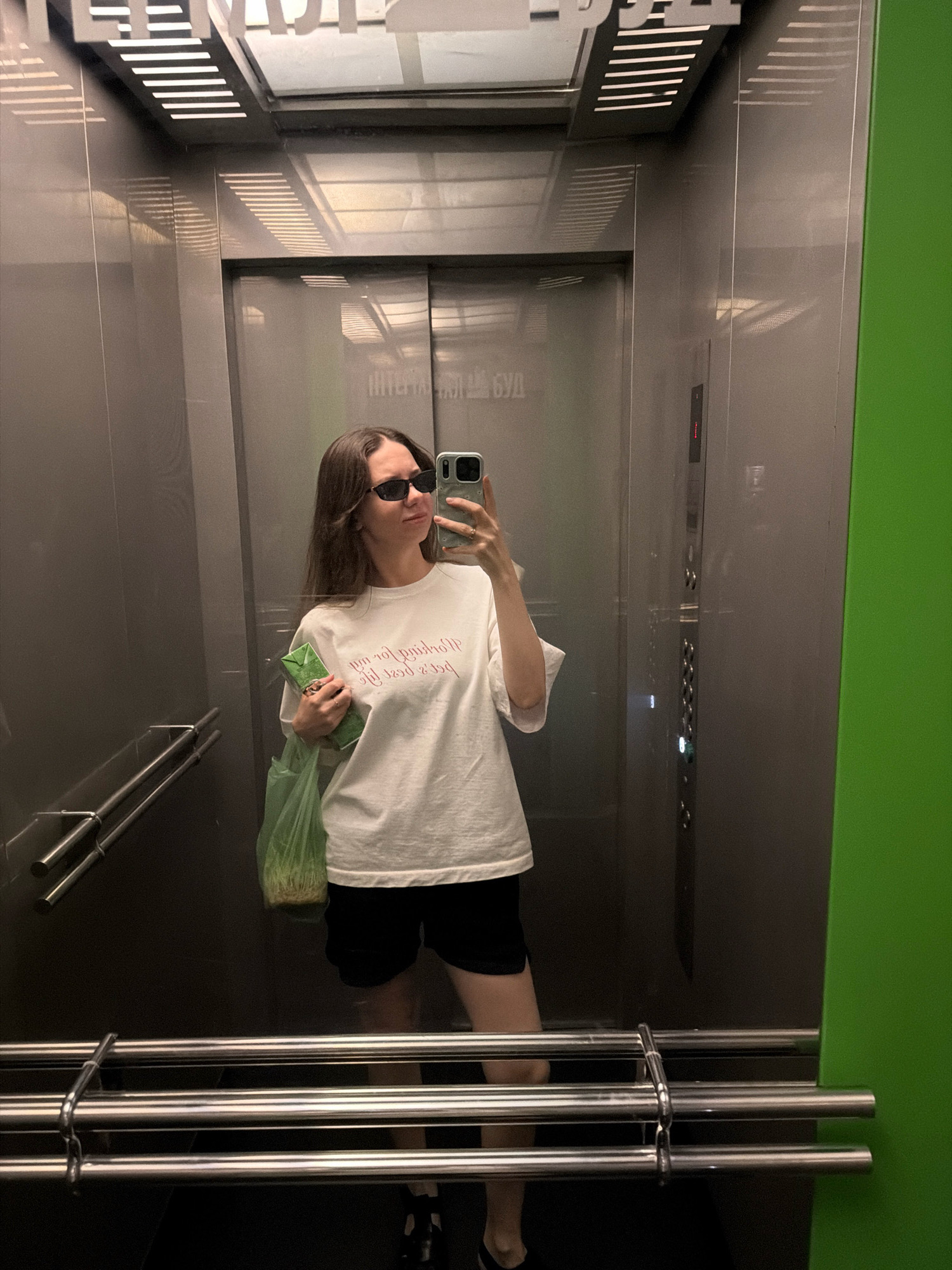 A young woman with long brown hair and sunglasses is taking a selfie in an elevator. She is wearing a white t-shirt with red text and black shorts. She is holding a green bag with a green carton inside in her left hand. The elevator has gray walls and metal railings. There is a bright green panel on the right side of the elevator. The text on the elevator walls appears to be in Cyrillic script. The scene is likely in Kyiv, Ukraine, given the Cyrillic writing and the prevalence of similar elevator designs in Ukrainian apartment buildings.