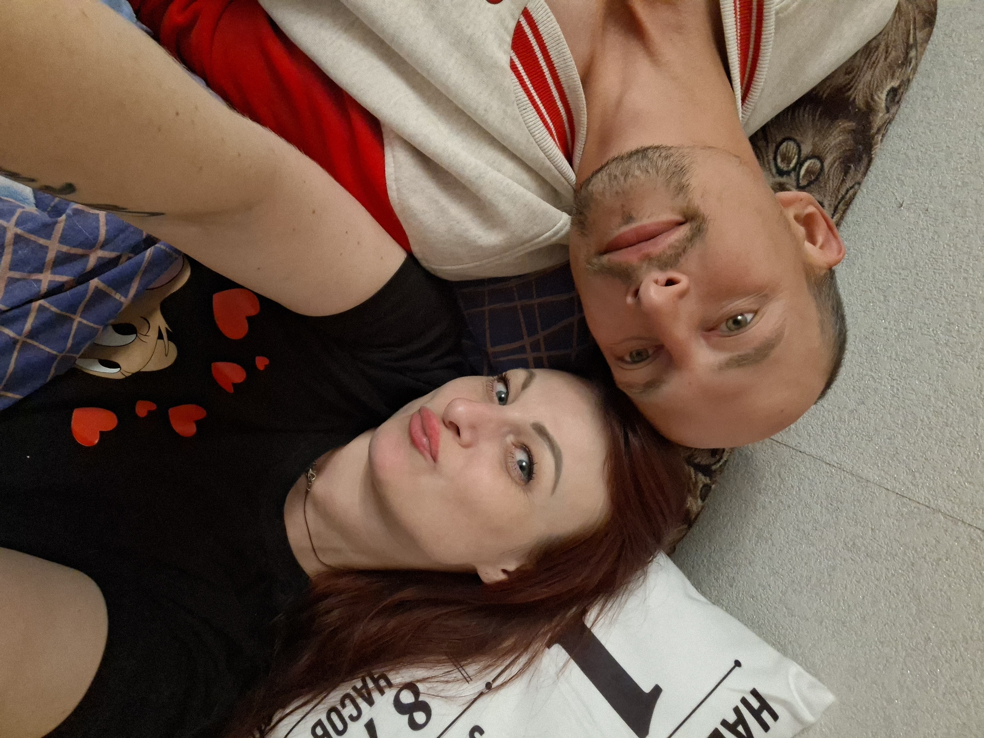 A close-up, high-angle indoor shot features a man and a woman lying down, looking up towards the camera. The image is oriented with their heads appearing upside down relative to the viewer. The woman, positioned in the lower half of the frame, has reddish-brown hair and light-colored eyes, and makes a slightly pursed-lip expression. She wears a black top adorned with several red heart shapes and a visible necklace. Part of her arm is visible on the left, resting on a blue and white patterned blanket which also shows a cartoon character's head (resembling Betty Boop).
The man is positioned above the woman, with his head resting near hers. He has short dark hair, light stubble on his face, and light-colored eyes, displaying a neutral, relaxed expression. He is wearing a grey and white striped jacket over a red garment. Behind him, a textured, light-colored surface and a darker patterned fabric are visible.
A white pillow with black text is seen at the bottom right of the frame, partially displaying words and numbers such as "JACOB," "8," and "HA". The scene depicts a casual and intimate moment between the two individuals, likely a selfie taken in an indoor home setting, within the region of Забалоцкі сельскі Савет, Belarus.