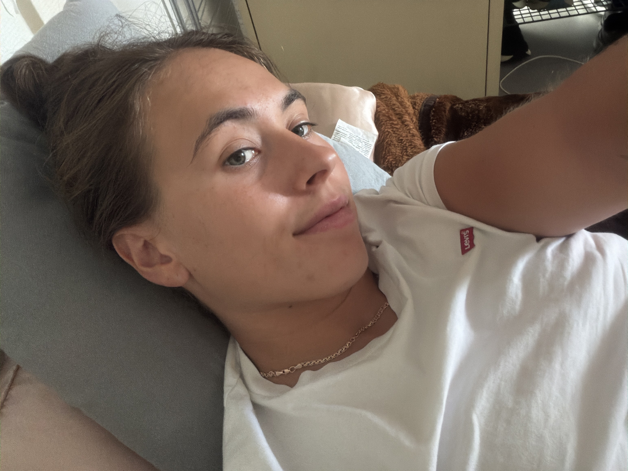 A young woman with brown hair pulled back in a messy bun is lying down, looking up at the camera. She is wearing a white t-shirt with a small red Levi's logo on the left chest. Her head is resting on a grey surface, and a brown furry blanket is visible behind her. The lighting suggests an indoor environment, possibly a bedroom or living area.