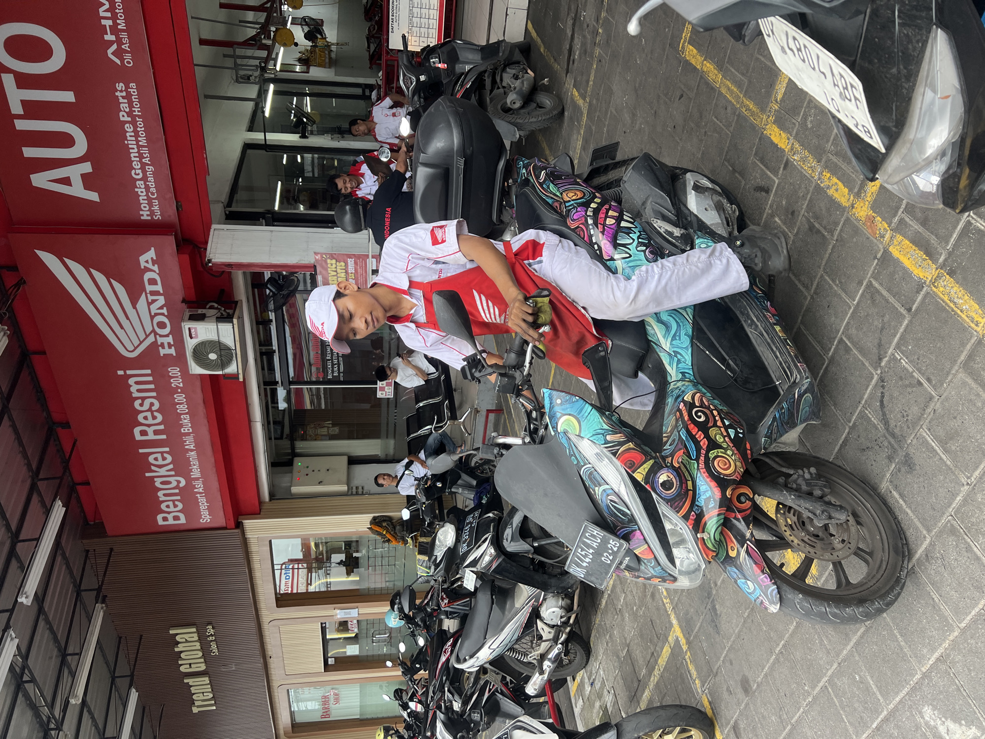 The image shows a scene in Kuta Selatan, Indonesia, likely at a motorcycle repair shop. A young man wearing a Honda uniform is sitting on a scooter with a distinctive colorful pattern, seemingly cleaning it with a cloth. Behind him is a storefront labeled "Bengkel Resmi Honda," indicating an authorized Honda repair shop. Other motorcycles are parked in the foreground. Two men in similar Honda uniforms are visible in the background inside the shop. A sign above the Honda shop reads "AUTO" and specifies that it sells Honda Genuine Parts. Adjacent to the Honda shop is a business called "Trend Global Salon & Spa." A license plate is visible on one of the motorcycles. The setting appears to be outdoors on a paved area.