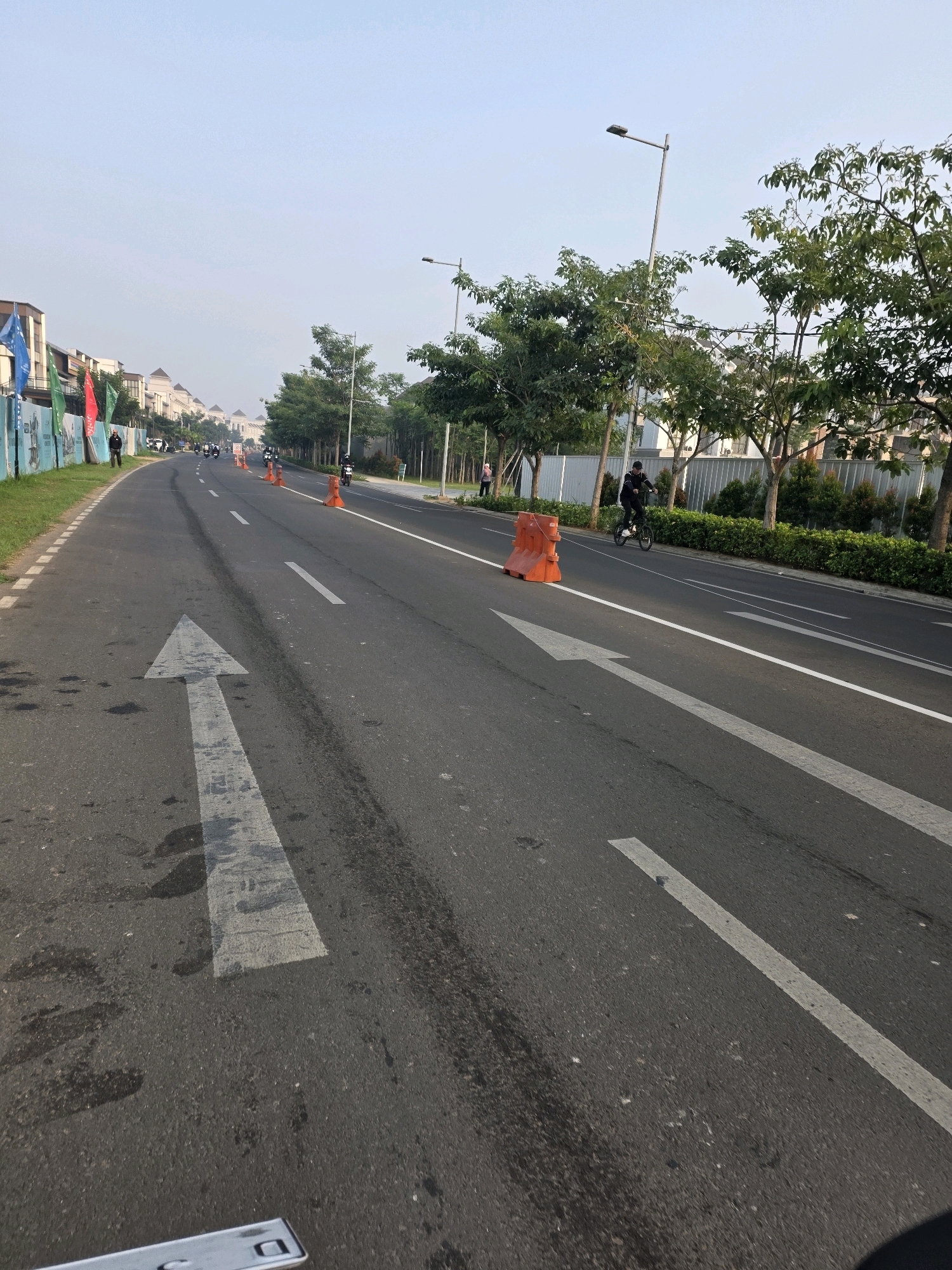 The image depicts a paved road with lane markings and directional arrows, likely in a residential or developing area of Bojongsoang, Indonesia. The road is lined with trees and some buildings are visible in the background. There are several orange traffic cones and a white plastic barrier on the side of the road. A person on a bicycle is visible riding on the road, and there are a few other individuals and vehicles in the distance, suggesting some pedestrian and vehicular activity. The sky is clear and the lighting suggests it is daytime.