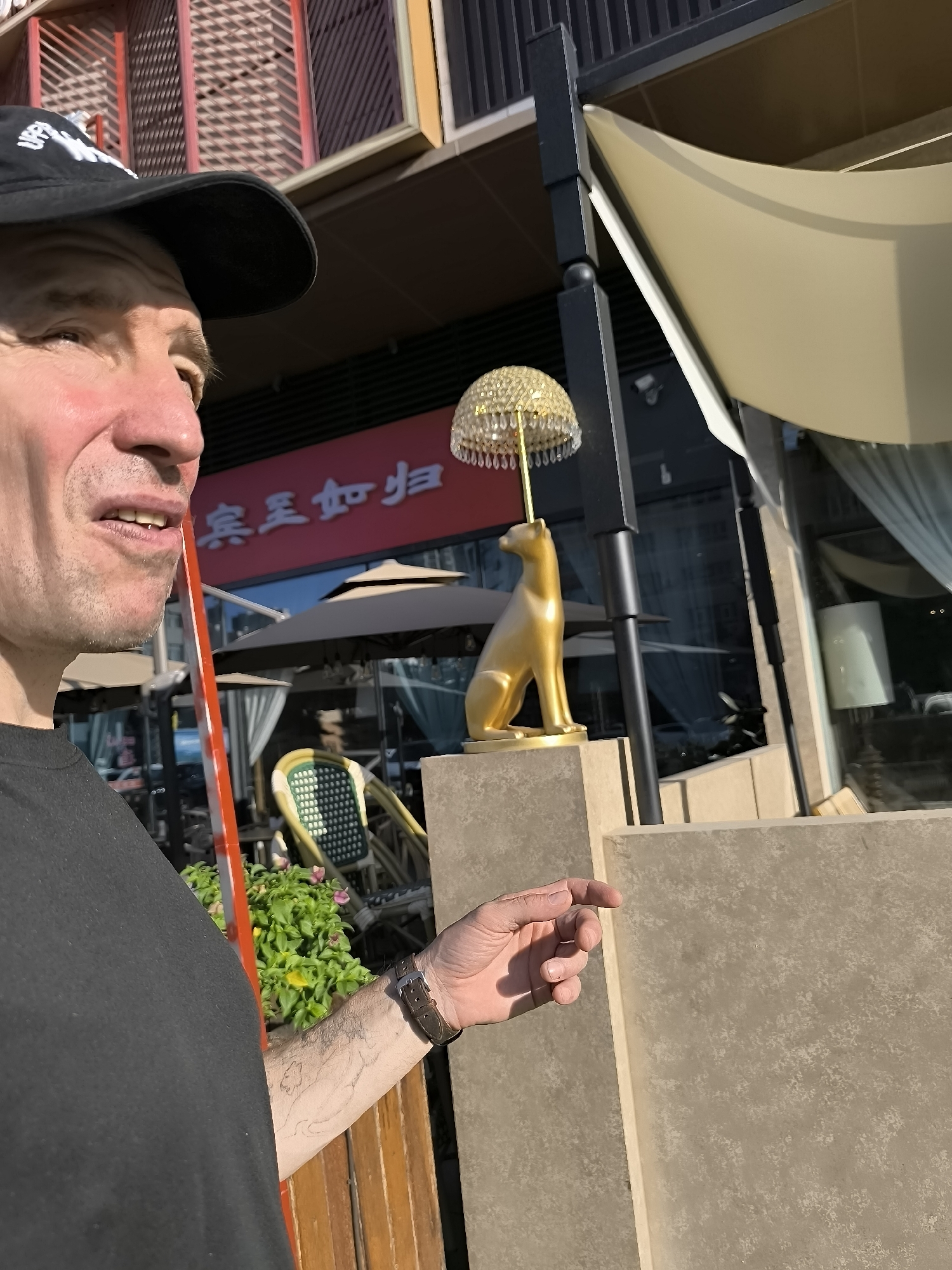A medium shot shows a man in a black baseball cap and black t-shirt on the left side of the frame. His face is turned to the right, and he is looking forward. He is holding a red straw to his mouth, which is slightly open. Behind him, there are buildings, including a building with red signage that appears to have Chinese characters. A gold statue of a seated dog is visible on a pedestal in the middle of the frame, topped with a bejeweled lampshade. The statue is to the right of the man's head. Further to the right, there is a cream-colored awning. The man's right hand is extended towards the right side of the frame, and he is wearing a watch on his wrist. The scene appears to be outdoors, possibly in a commercial or dining area with tables and chairs visible in the background. The lighting suggests it is daytime.