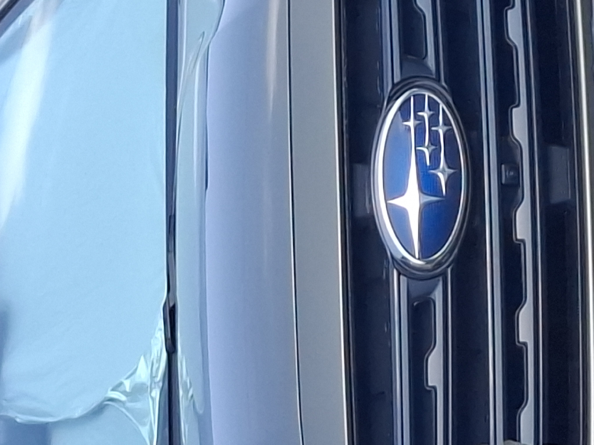 An extreme close-up shot focuses on the front grille and emblem of a dark-colored Subaru vehicle. The emblem, a blue oval with a starburst design, is prominently displayed. The grille features horizontal slats with a textured pattern. The upper portion of the image shows the hood of the car and a light blue sunshade that is partially covering the windshield. The scene appears to be outdoors, likely in a city environment like Almaty, Kazakhstan, given the context, but the specific location within Almaty cannot be determined from the image alone. There are no people or any observable actions taking place. The lighting suggests it is daytime, with soft, diffused light.