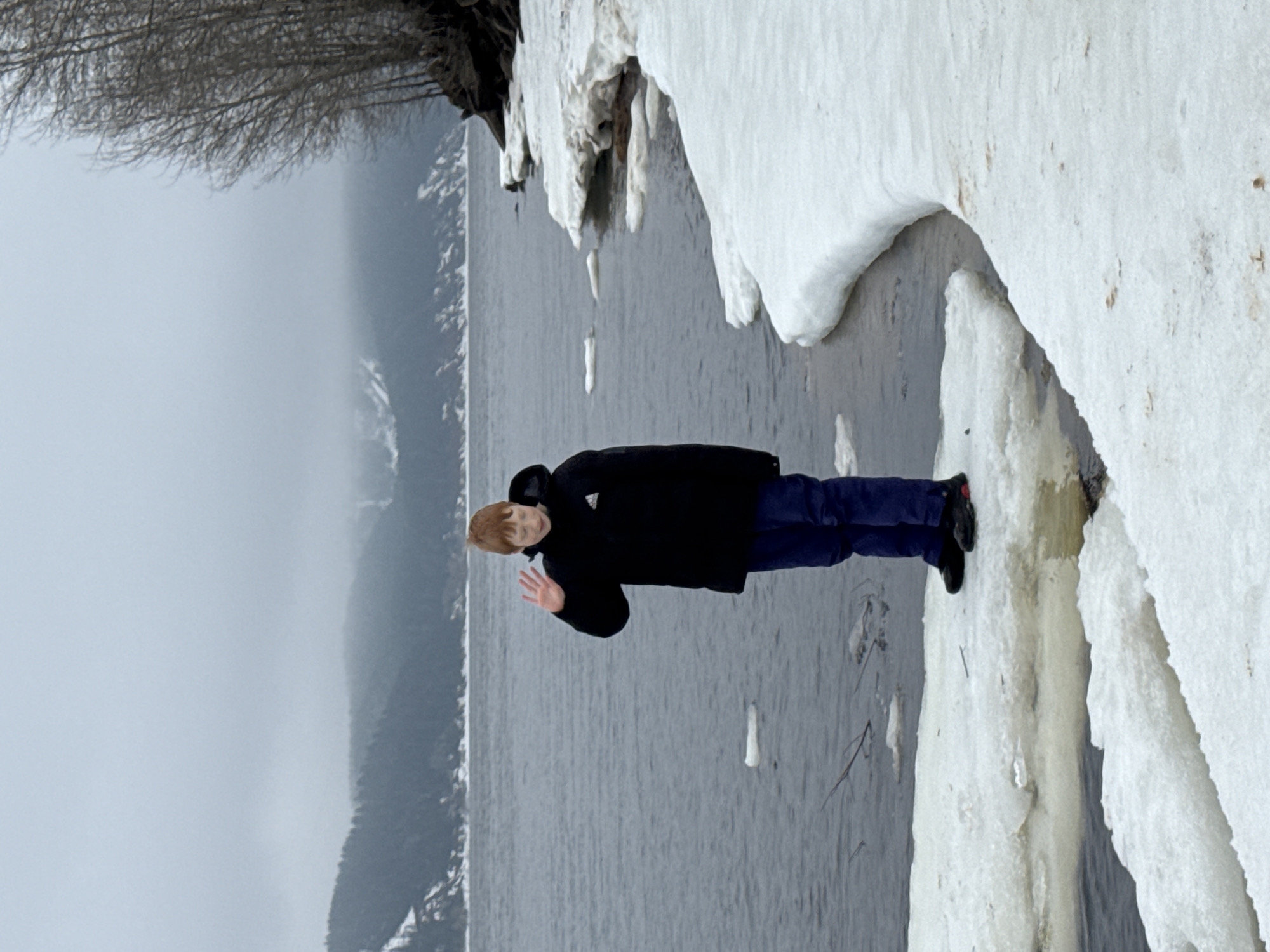 The image shows a person standing on a snowy bank beside the Kama Reservoir in Chaikovsky, Russia. The person, wearing a black jacket and blue pants, is waving. The reservoir is visible in the background, with a forest on the opposite shore. The snowy bank is covered in thick ice formations. The sky is overcast and gray.