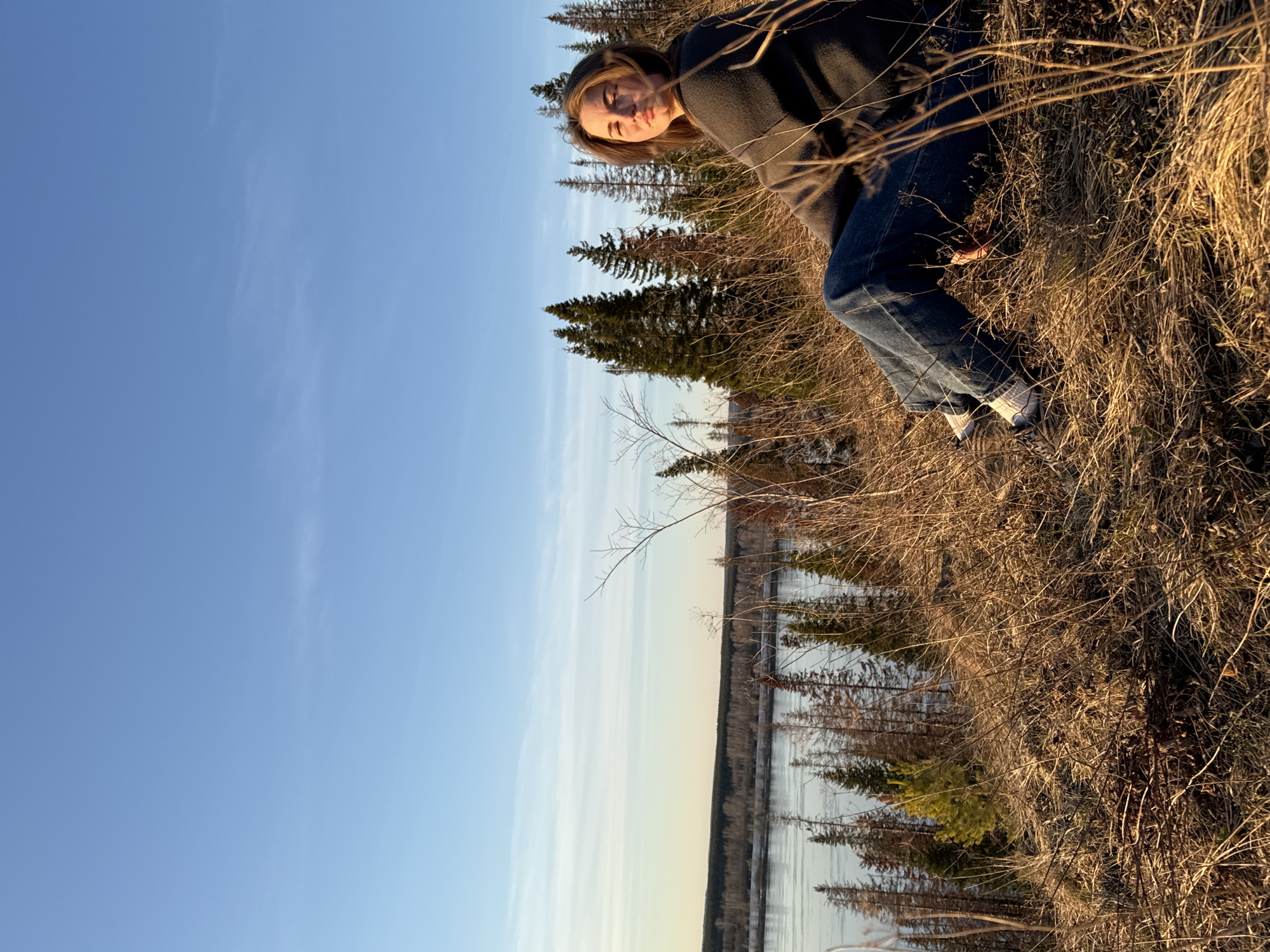 The image depicts a young woman standing on a dry, grassy bank in what appears to be a natural setting. The woman is wearing a dark sweater, blue jeans, and light-colored sneakers. Behind her are coniferous trees, and in the distance, a body of water is visible. The sky is blue with some wispy clouds. The location appears to be Ольховка, Russia.