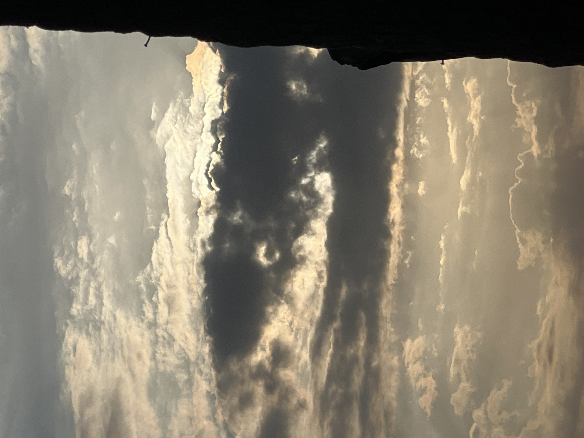 A silhouette of a dark structure, possibly a building, is visible on the right side of the frame. The structure is dark and angular, with a few small, lighter-colored protrusions along its edge. The rest of the image is dominated by a cloudy sky. The clouds are varied in density and color, ranging from dark gray to bright white and tinged with yellow and orange, suggesting either sunrise or sunset. Some clouds are thin and wispy, while others are thick and billowy. The overall impression is of a dramatic sky over Kuta, Indonesia, a popular tourist destination known for its beaches and sunsets.