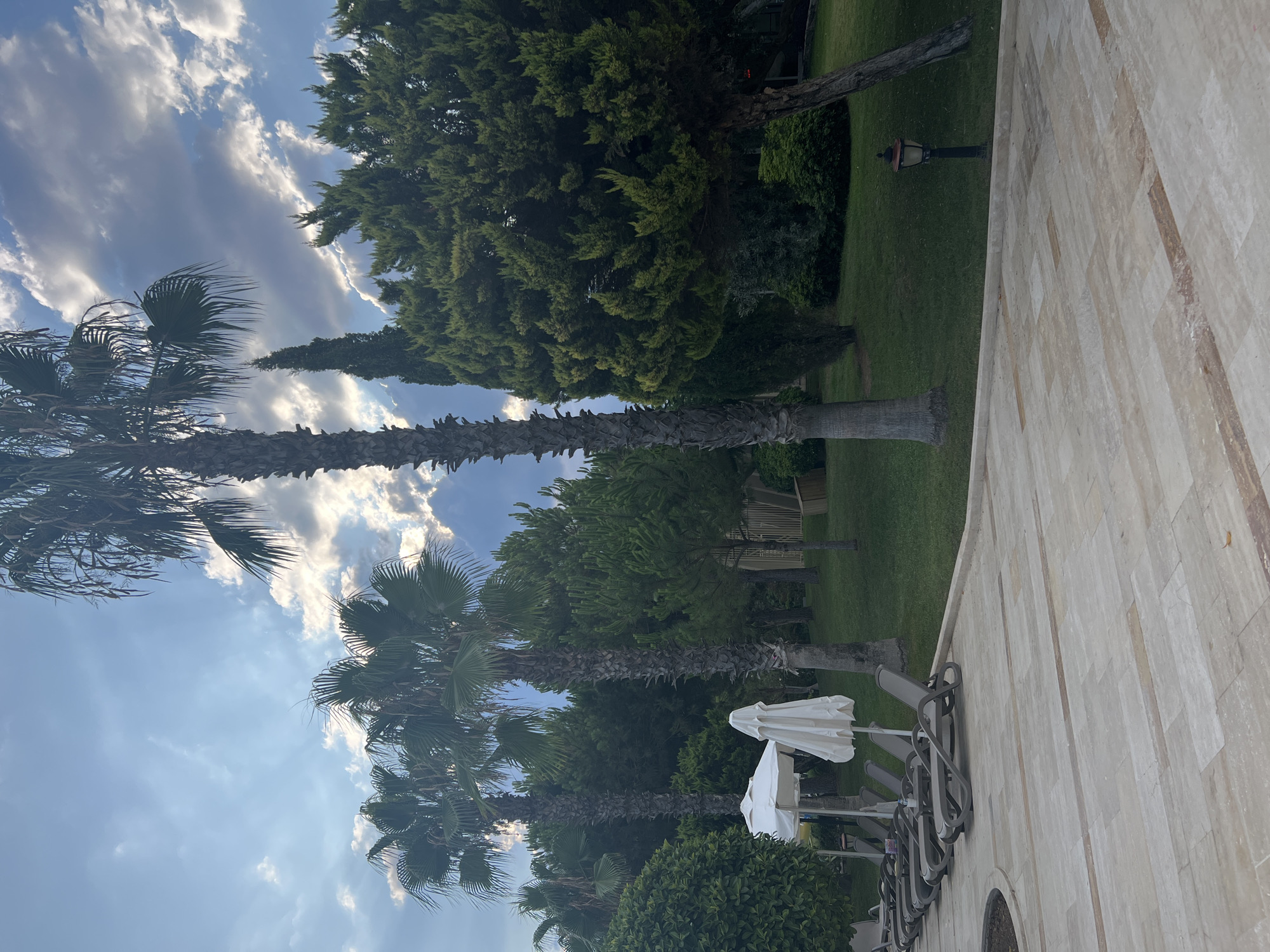 This image depicts an outdoor scene likely within a resort in Serik, Turkey. In the foreground, a paved area with large, light-colored rectangular tiles leads towards a manicured lawn. A line of dark gray lounge chairs with white umbrellas stands on the edge of the lawn. Towering palm trees and dense evergreen trees dominate the middle ground, creating a lush, tropical atmosphere. The sky above is a mix of blue and dramatic clouds, with sunlight filtering through in some areas. A dark, antique-style lamp post is visible on the right side of the lawn. The overall impression is one of a peaceful, well-maintained outdoor recreational space.