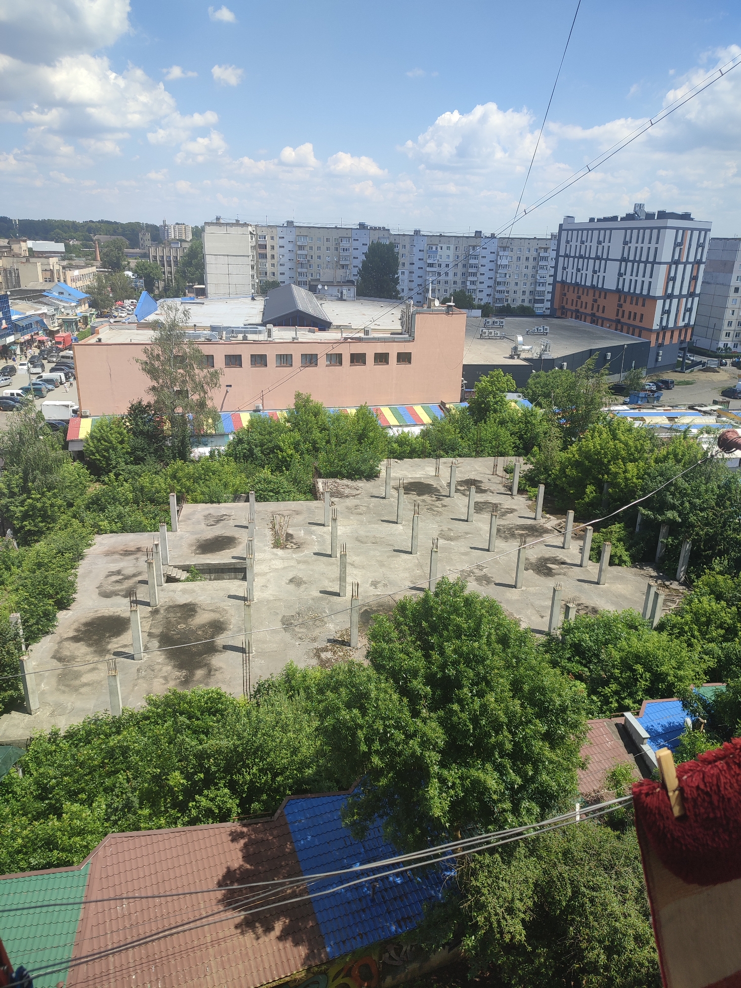 This is an aerial, eye-level shot of an urban landscape in Chernivtsi, Ukraine. The foreground features the roofs of two buildings, one with a red tiled roof and another with a blue tiled roof, both showing signs of wear. A red towel hangs from a clothesline, secured with a wooden clothespin. Further into the image, an unfinished concrete structure with numerous pillars stands amidst lush green trees. Beyond this, a light pink, rectangular building with dark-trimmed windows is visible, featuring a colorful striped awning along its lower facade. In the background, a dense arrangement of apartment buildings, characterized by various architectural styles and colors, stretches across the horizon, under a bright blue sky dotted with fluffy white clouds. The scene captures a mixture of ongoing construction, established residential areas, and abundant greenery.