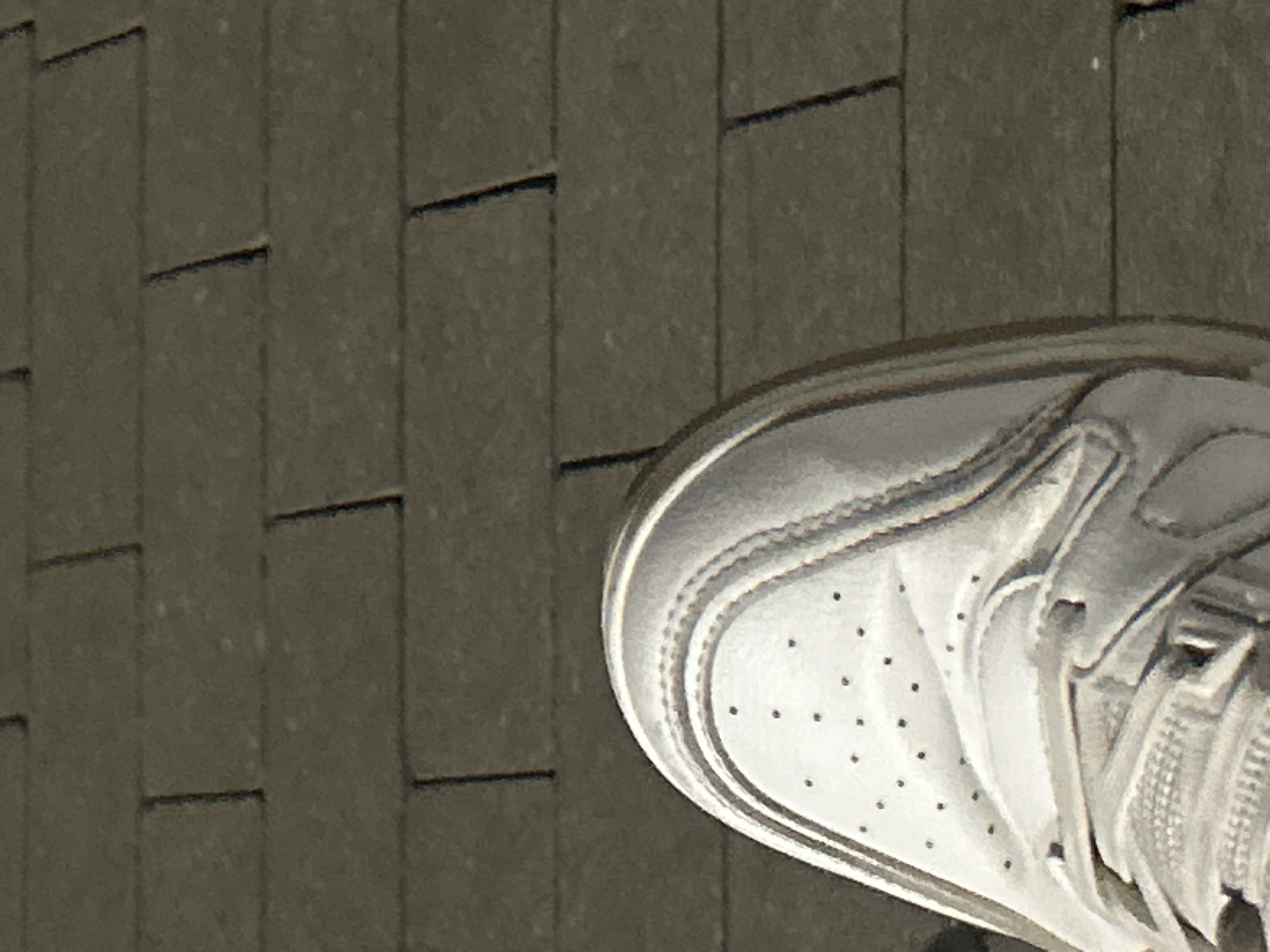 The image shows a close-up view of the toe area of a white sneaker resting on a textured, dark grey surface. The surface appears to be made of rectangular paving stones or tiles, arranged in a grid pattern with visible grout lines. The sneaker is white with perforations on the toe box, characteristic of athletic footwear. Stitching details are visible along the seams of the shoe, and the sole has a raised tread pattern. The lighting suggests it is daytime, with shadows casting from the shoe onto the paving stones, indicating a sunny or brightly lit environment. The perspective is from a high angle, looking down at the shoe and the ground. There is no visible text in the image. The setting appears to be outdoors, possibly a street, sidewalk, or patio in Siedlce, Poland. No specific activity or event is depicted, but the image focuses on the shoe and its immediate surroundings, suggesting a moment of pause or a detail shot.