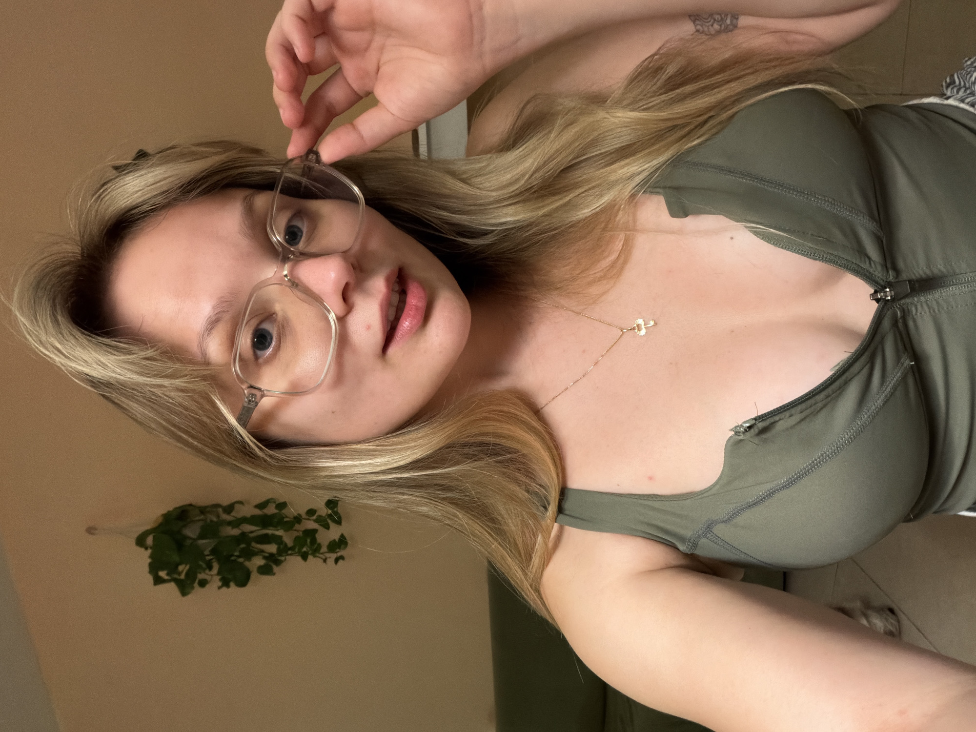 A blonde-haired woman wearing glasses is the primary subject of the image. She is also wearing a green, zip-up top and a delicate gold necklace with a small pendant. A tattoo is visible on her upper arm. She is holding her glasses in her right hand. In the background, there is a plant hanging on a light beige wall. No information suggests this picture was taken in Tel-Aviv, Israel.