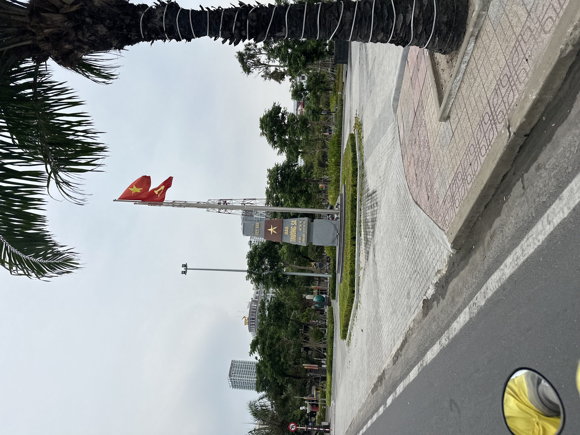Captured at an oblique angle, the scene shows a public space in the city of Nha Trang Ward, Vietnam, with a prominent sign, partially obscured by trees. The sign is inscribed with the words "CHXHCN VIET NAM" and "ĐẢO TRƯỜNG SA" along with geographical coordinates. Vietnamese flags atop a metal structure flutter in the wind. Lush trees line the area behind the sign and along the sidewalk. A patterned sidewalk is visible next to the road. A fragment of a palm tree frames the upper left corner of the image. The asphalt road is in the foreground. A blurred motorcycle mirror is visible at the lower right of the image. Buildings rise in the distance. The sky is overcast.