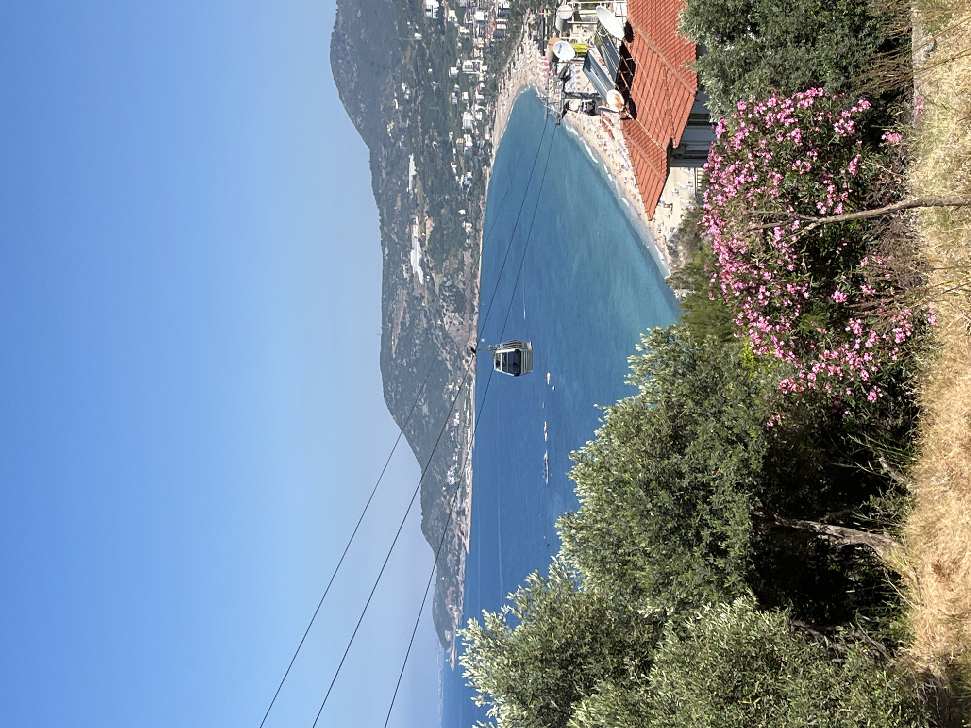 The image showcases a cable car traversing the landscape in Alanya, Turkey. The cable car, gray in color, is suspended mid-air, connected to its cables. The scene is partially framed by green trees and flowering bushes with pink blooms. Beyond the cable car, there is a panoramic view of the turquoise sea meeting a sandy beach crowded with people. Buildings are situated along the coastline. In the background is a mountain. The sky is a clear blue.