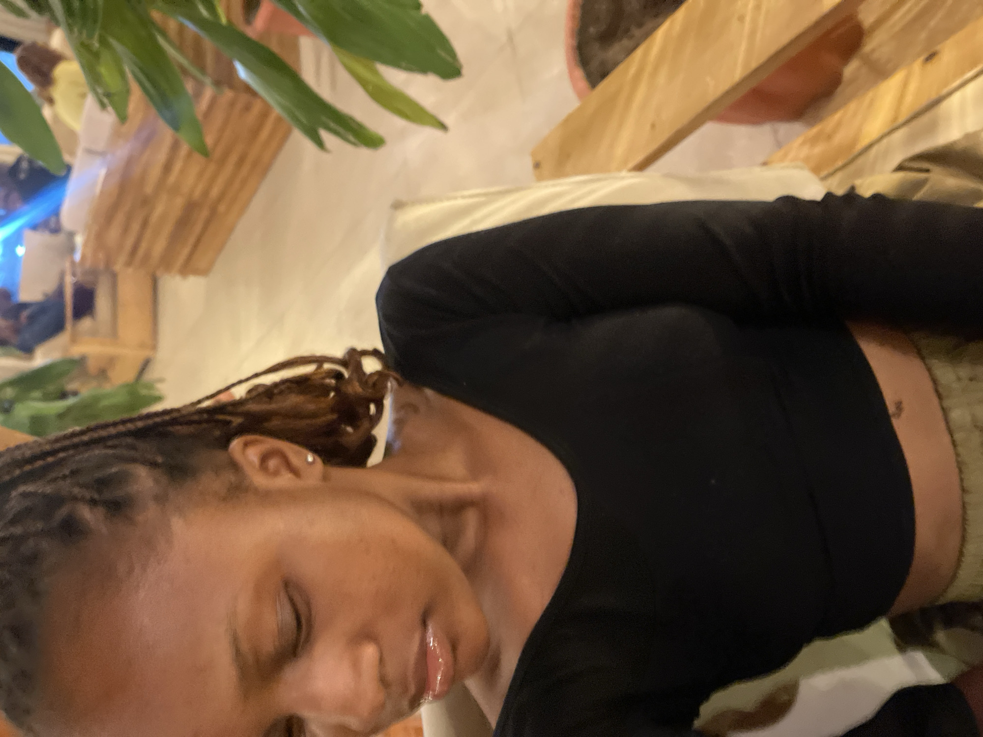 The media file shows a close-up, rotated view of a young Black woman with braided hair. She is wearing a black long-sleeved top and patterned pants. Her eyes are closed, and her head is tilted down, giving a relaxed or contemplative appearance. She has a silver stud earring in her left ear.
The background suggests an indoor setting, possibly a restaurant or cafe, with wooden furniture and greenery visible. The lighting appears to be artificial, indicating it could be evening or nighttime. The overall ambiance is casual and relaxed. There is no visible text in the image. The location context provided is Gura Top, Nigeria.