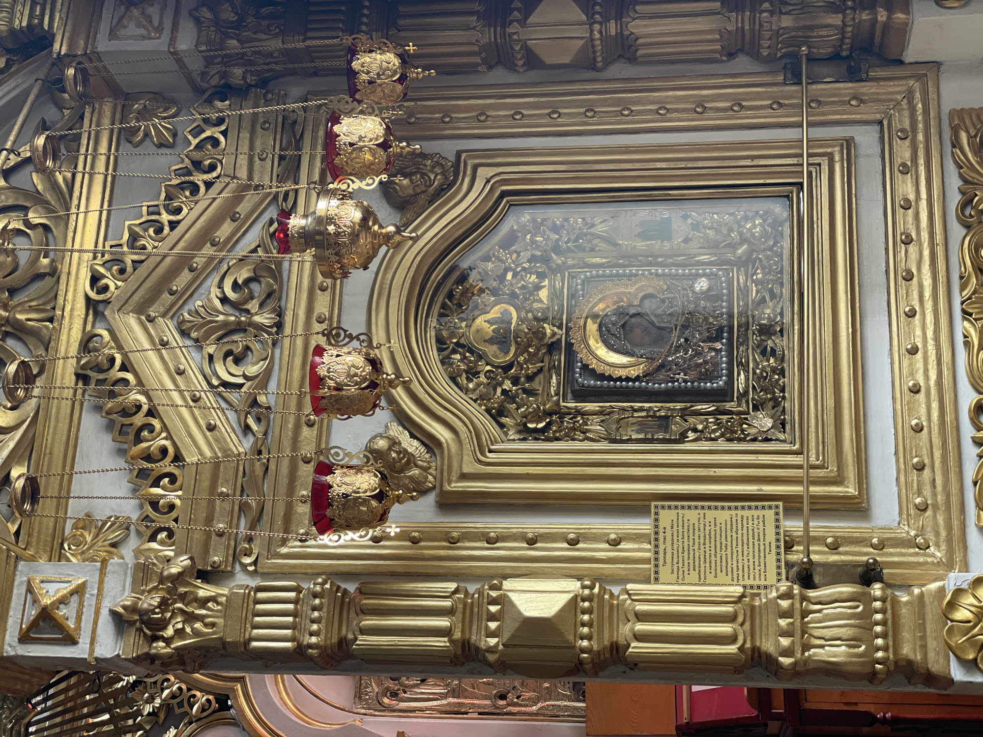 A detailed view of a gilded and ornate religious object, likely an icon or shrine, is presented. The object features a frame with intricate carvings and a small, framed image, partially obscured, under glass. A small, framed information placard, containing text in Cyrillic, is attached beneath the object. Four hanging, decorative lamps or censers with red accents are suspended via chains to the left of the frame. The environment is a religious setting, likely located in the city of Kazan, Russia.