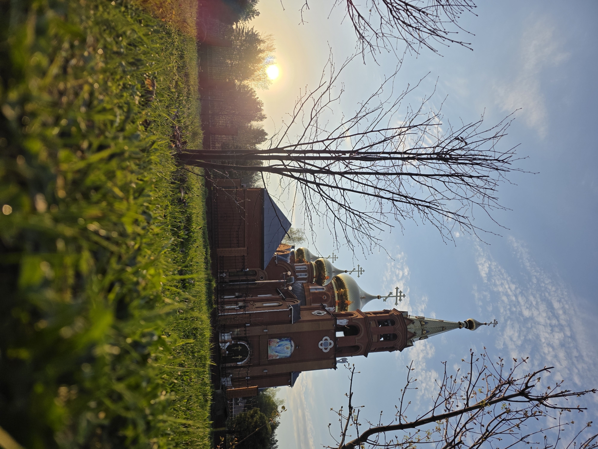 The image shows the Holy New Martyrs and Confessors of Russia Church in Chaikovsky, Russia, viewed from a low angle. The brick church features golden domes and a tall spire, standing against a bright blue sky with scattered clouds. Bare tree branches frame the structure. In the background, a wooded area is visible, with the sun setting behind the trees. Grass grows along the foreground, suggesting a natural setting. The overall impression is one of serene spirituality.