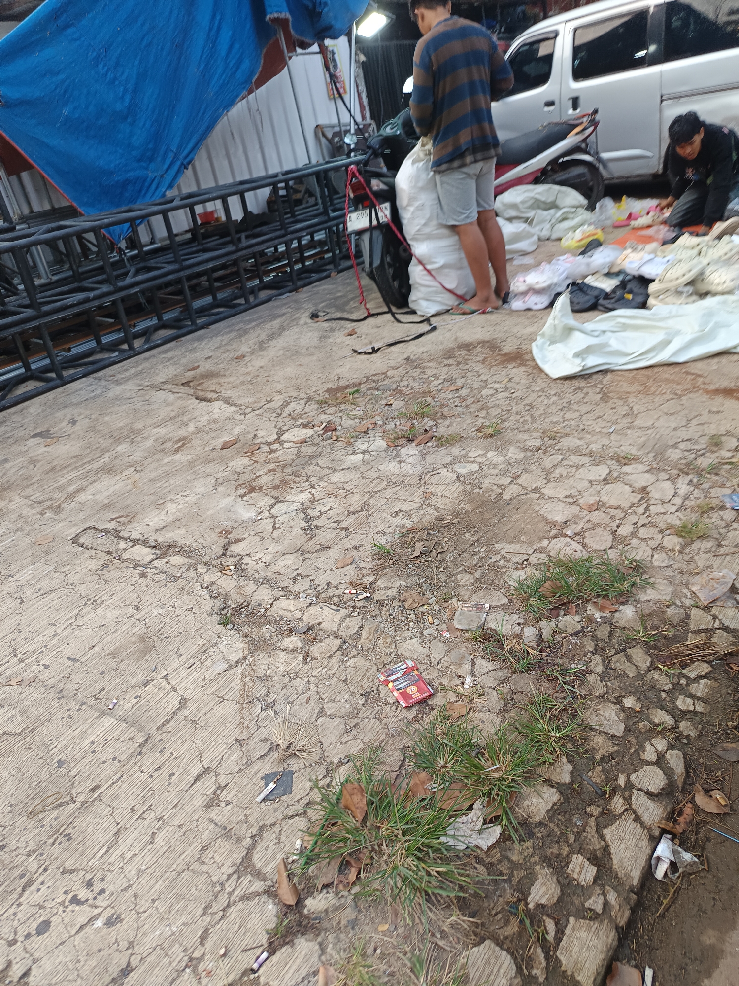 An outdoor scene depicts a cracked concrete ground with patches of dry grass and scattered debris. In the background, a silver van and a motorcycle are visible. A person is seen crouching and handling various items, likely clothing, spread out on the ground. Another person stands near the motorcycle, seemingly engaged in a similar activity. A large blue tarp is draped over some metal structures in the upper left portion of the image. The overall environment suggests an area in Sodong, Indonesia, possibly a marketplace or a storage area.
