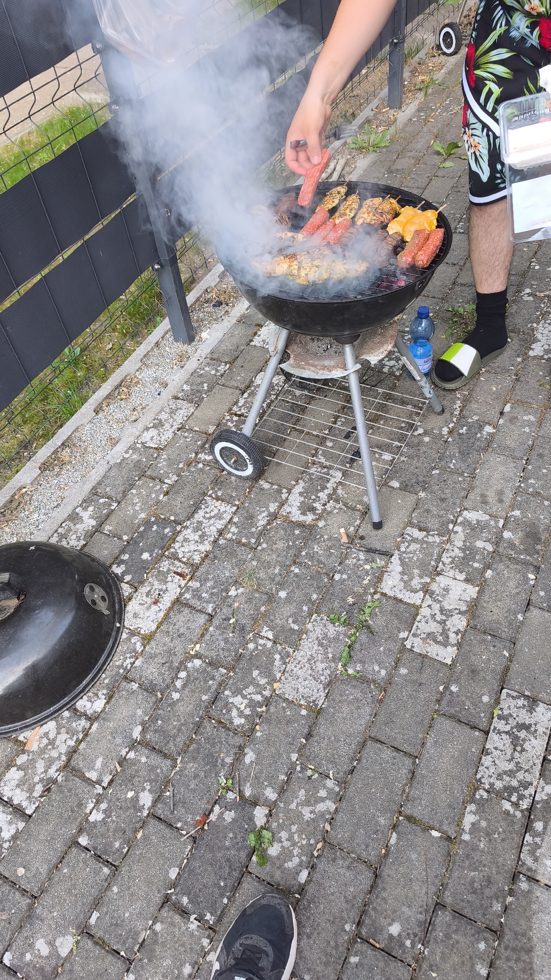 The image depicts an outdoor grilling scene on a paved surface in an unknown, unknown city. In the upper-center, a black, circular charcoal kettle grill emits a substantial amount of white smoke. On the grill grate, various food items are cooking, including several reddish-pink cylindrical items (appearing to be sausages or ground meat skewers), multiple skewers with cubed, yellowish meat (likely chicken), and some cut pieces of golden-yellow produce (possibly bell peppers).
A person's bare right arm and hand are visible on the right side of the grill, holding a pair of tongs, actively placing or turning one of the reddish-pink items. This individual is wearing black shorts with a tropical pattern featuring green leaves and red floral elements, black socks, and a white slide sandal with a neon green stripe on their right foot. Near the grill's base, a small blue plastic water bottle and a small blue/grey canister are situated on the grey paver stones. A clear plastic container with a lid is held in the person's left hand, partially visible near their right hip.
To the bottom-left of the frame, a black, domed grill lid rests on the paved ground. The ground surface consists of rectangular grey paver stones, which exhibit various patches of lighter discoloration, possibly moss, lichen, or efflorescence, along with small bits of debris and sparse green vegetation growing in the cracks. In the bottom-center foreground, a left foot wearing a black and white athletic shoe (with a visible "Nike" logo) is partially visible.
In the background, a black metal mesh fence with horizontal privacy elements separates the paved area from an expanse of green grass and a strip of gravel or dirt. A small white wheel from an unknown object is partially visible behind the person's shorts, near the fence line.