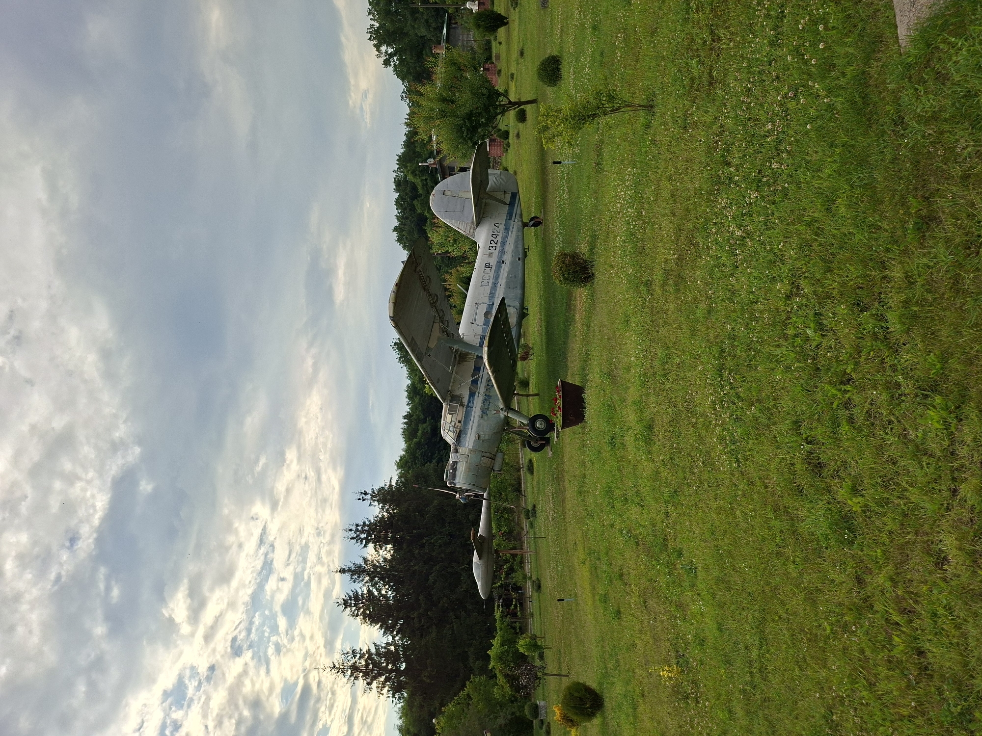 The image depicts an outdoor scene in Malcoci, Moldova, featuring a vintage Antonov An-2 biplane parked on a grassy lawn. The plane is painted light blue and white, with visible markings that include "CCCP 32424". It is positioned on its landing gear. The surrounding environment is a well-maintained park or garden with lush green grass dotted with small white flowers. Several trees, including evergreen conifers and deciduous trees, are visible in the background, along with a few bushes and small ornamental trees in the foreground. In the distance, part of a building with a red brick chimney can be seen, suggesting the presence of structures nearby. The sky is overcast with a mix of light and dark clouds. The overall impression is that of an open-air display or a historical artifact preserved in a tranquil setting.