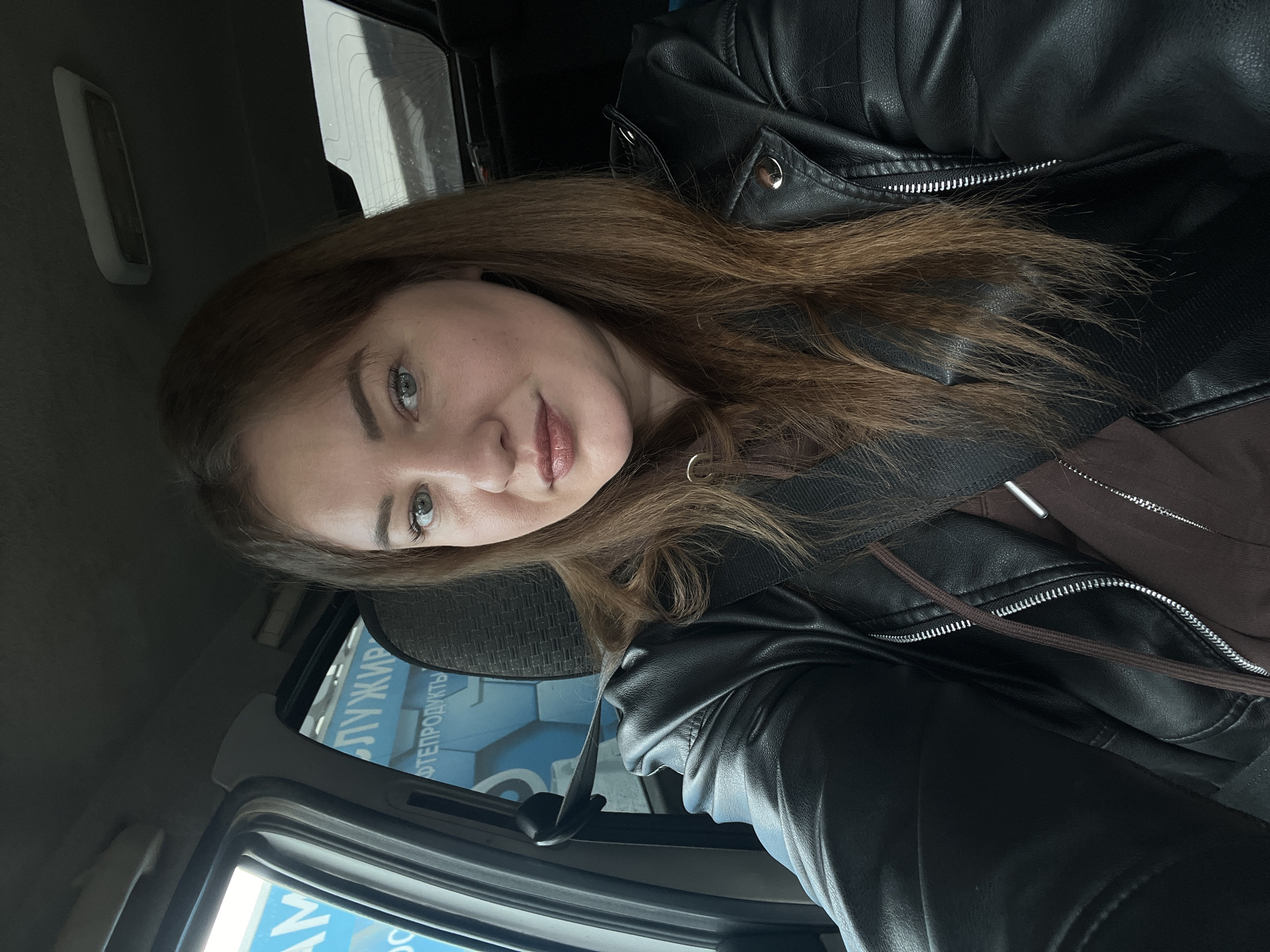The image shows a woman with long brown hair, wearing a black leather jacket and a brown hoodie, inside a car. Her eyes are light colored. Through the car window, part of a building with signage in Russian, including the words "ЛУЖИР" and "ПРОДУКТЫ", can be seen. Based on the Russian text, the location is likely in Russia. With additional knowledge, it can be determined this location is in the city of Chaikovsky, Russia. The woman is looking at the camera with a slight smile. The scene suggests a casual moment inside a vehicle.