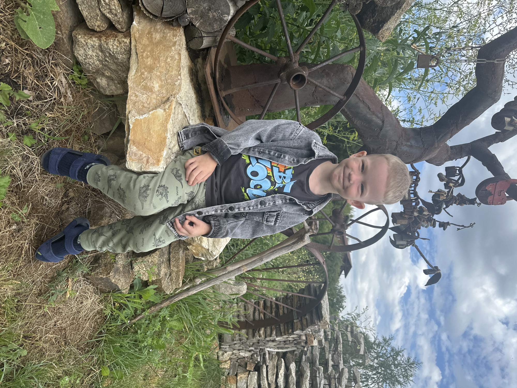 A young boy with blonde hair and blue eyes is sitting on a stone bench outdoors. He is wearing a grey denim jacket over a black t-shirt with colorful lettering and green pants with a dinosaur pattern. The boy is smiling at the camera. Behind him, there is a large, rusty metal sculpture resembling a tree with locks hanging from its branches. To his left, a large wagon wheel is visible, and to his right, a stack of flat stones forms a wall. The environment appears to be rural, with green grass, vegetation, and a cloudy sky overhead. The location is specified as Danilovka, Altai Krai, Russia.