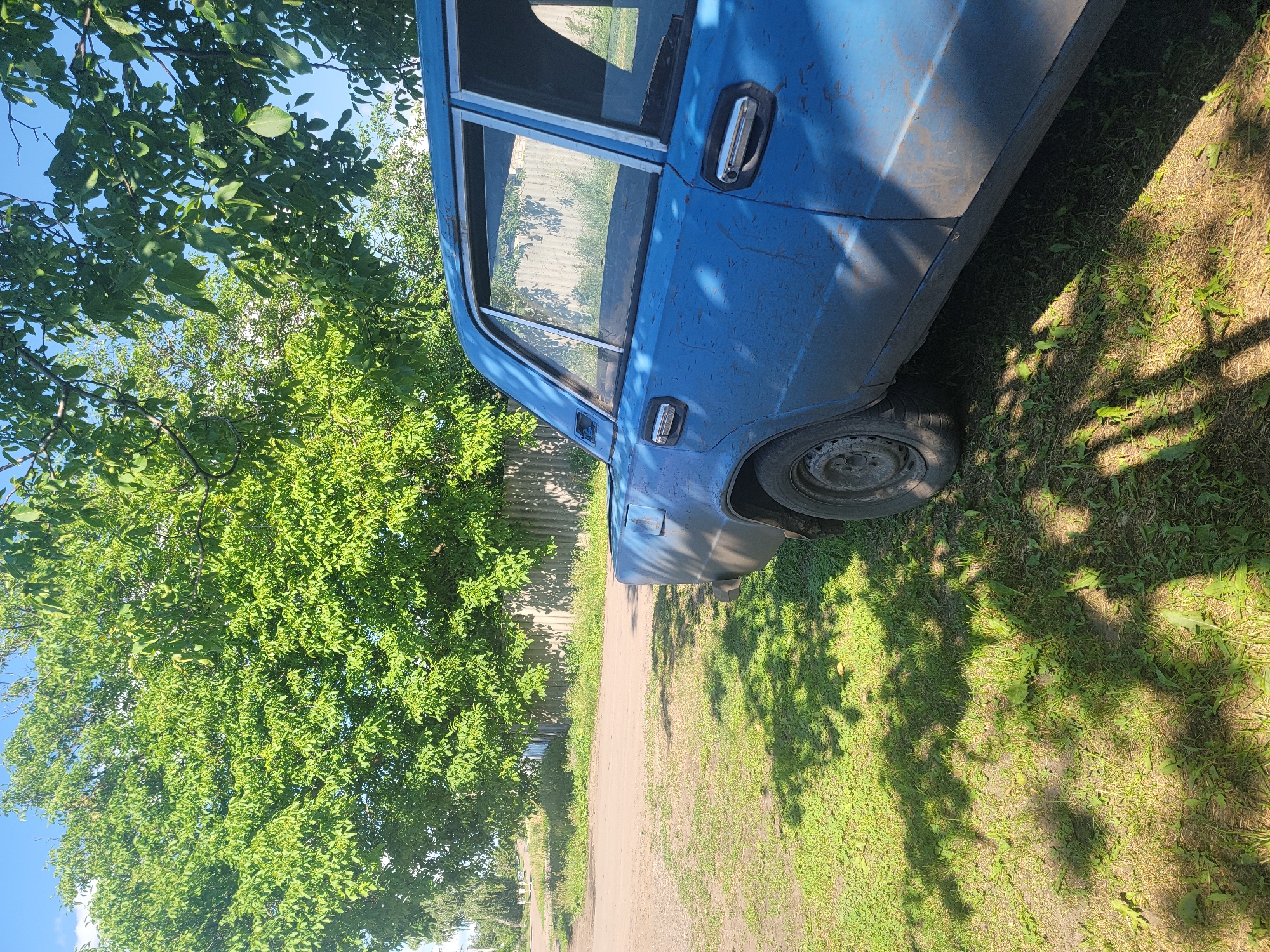 The image shows a blue, weathered car parked on a patch of grass next to a dirt road in the village of Vovkivka, Ukraine. The car, an older model, is partially obscured by shadows cast from overhead trees, suggesting a sunny day. To the left of the car, the dirt road continues and appears to lead to a residential area with a fence visible in the background. Lush green trees with dense foliage dominate the upper portion of the image, with the sky peeking through the leaves. The grass is green and somewhat patchy, with some dried leaves and debris scattered on the ground. No people are visible in the scene.