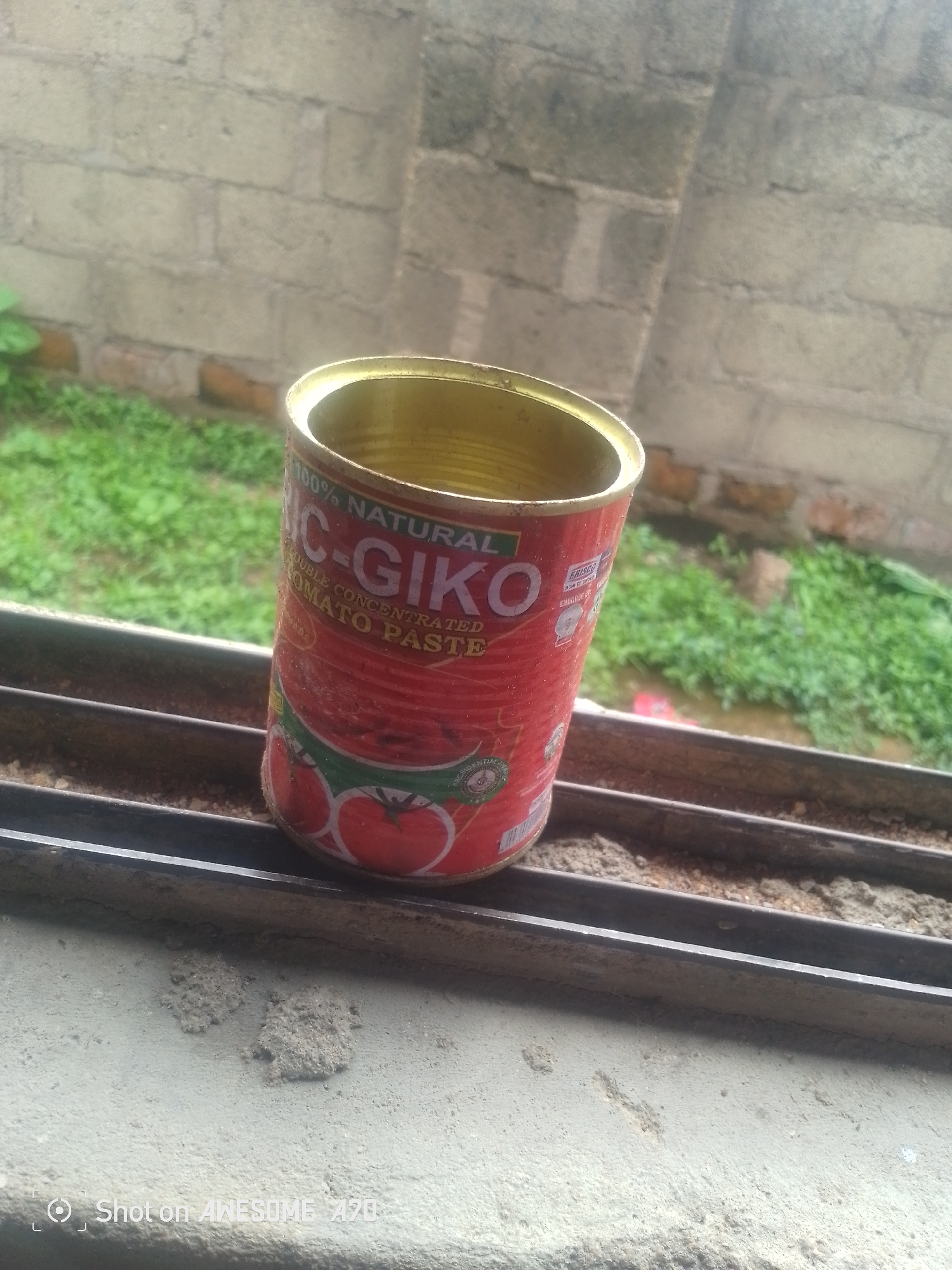 A close-up, angled shot shows a red, empty tin of tomato paste. The tin is positioned on a metal ledge or railing, with a brick wall and green foliage in the background. The label on the tin is partially visible, reading "100% Natural MC-GIKO DOUBLE CONCENTRATED TOMATO PASTE." The lid of the tin is open, revealing the empty interior. The foreground shows a rough concrete surface with two small, clumpy deposits of what appears to be dirt or sand. A watermark at the bottom left corner of the image reads "Shot on AWESOME A70."