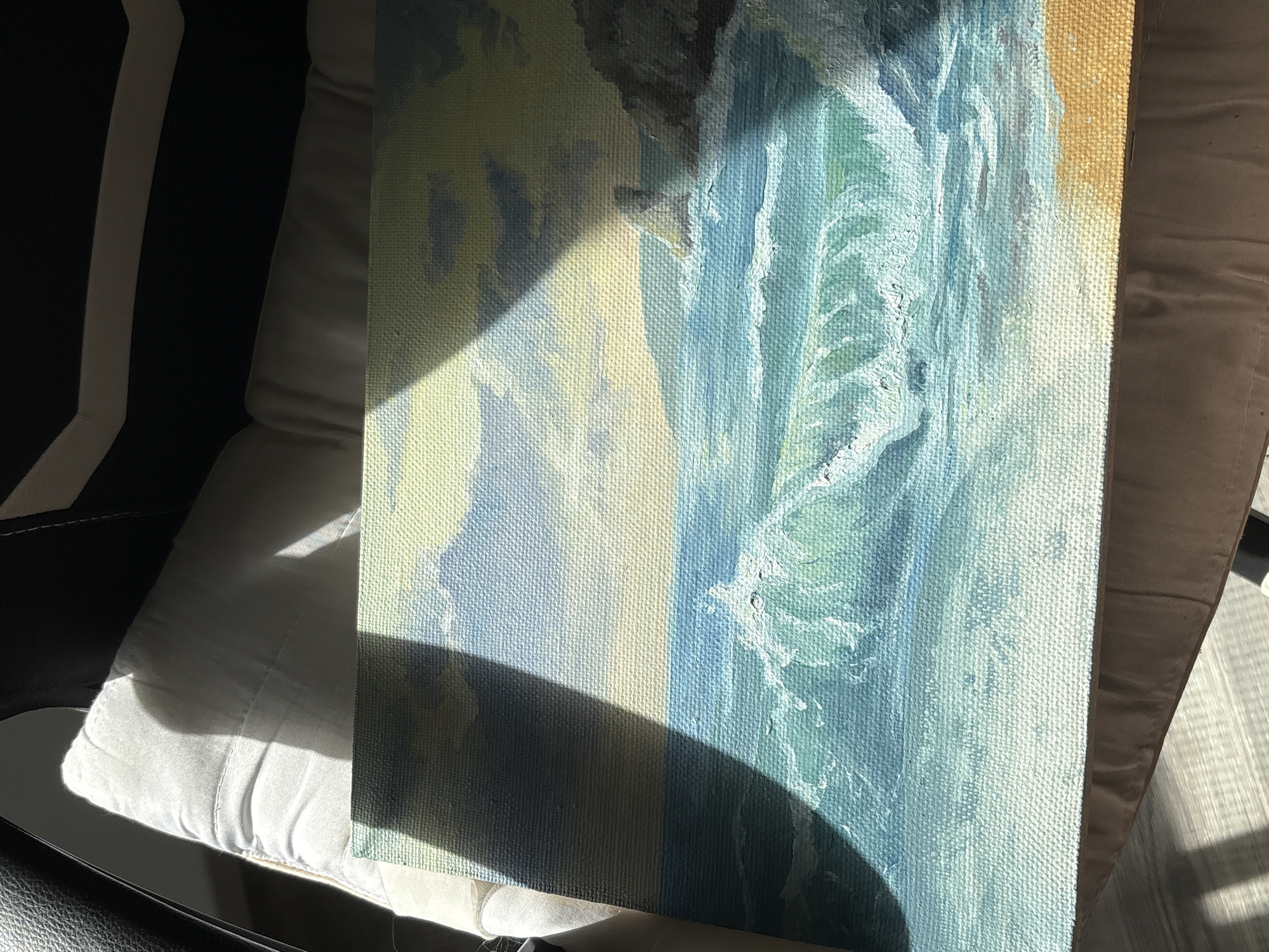 The image shows a partially lit canvas resting on a light-colored cushion, possibly part of a chair. The canvas features a painting with a coastal theme, showcasing cliffs, waves, and sand. The painting employs varying shades of blue, green, yellow, and brown. A dark shadow partially obscures the canvas, suggesting a nearby object blocking the light. The background includes parts of a black and white chair. There's no explicit evidence to suggest that this image or painting is associated with the city of Saint Petersburg, Russia. The elements of the scene are indicative of an indoor setting with artistic elements.