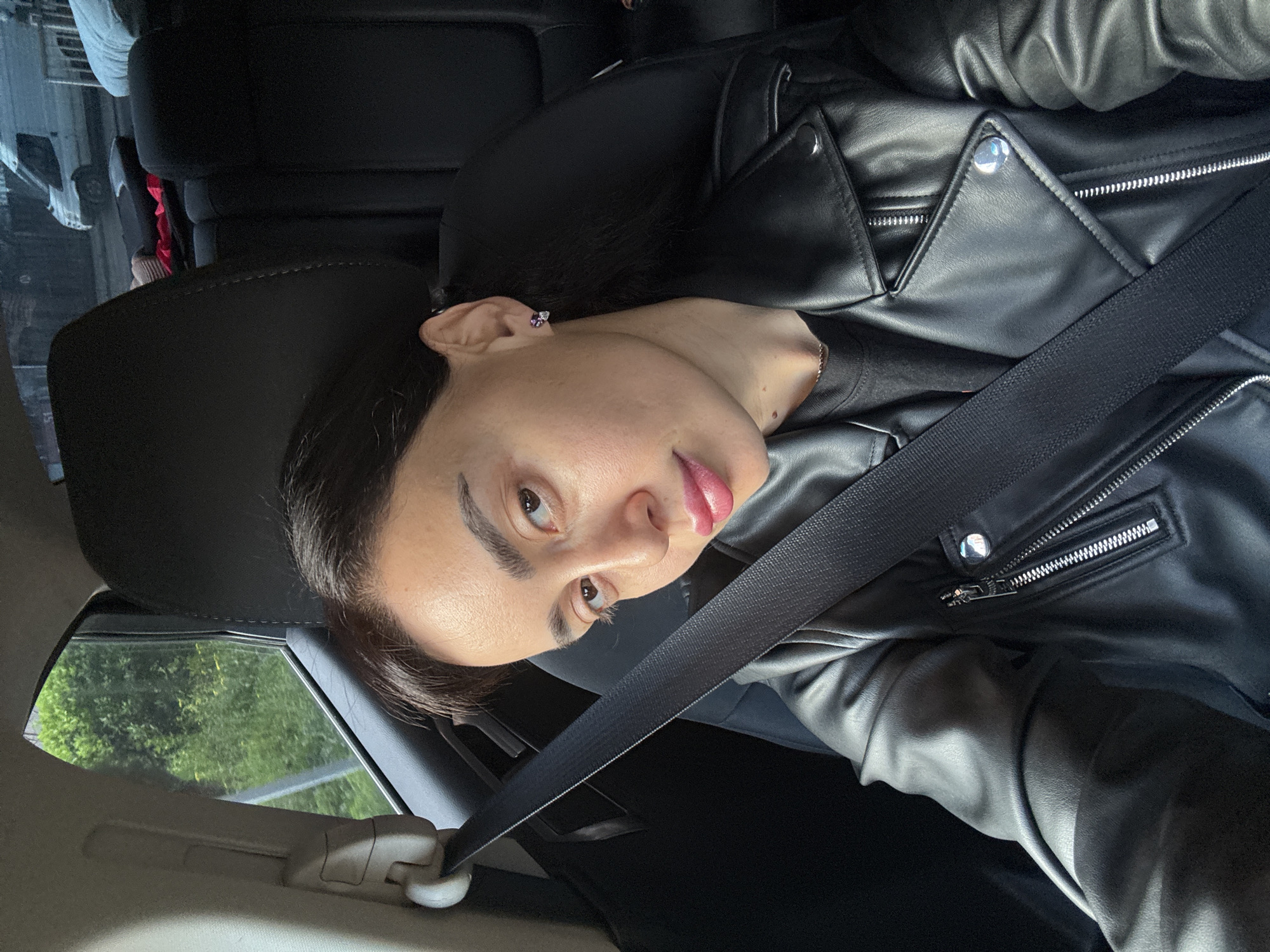 A woman with brown hair is in a car, wearing a black leather jacket and a seatbelt. She has light skin and is wearing a small purple earring. The car appears to be moving, as green trees are visible through the window. The location cannot be determined from the provided information.