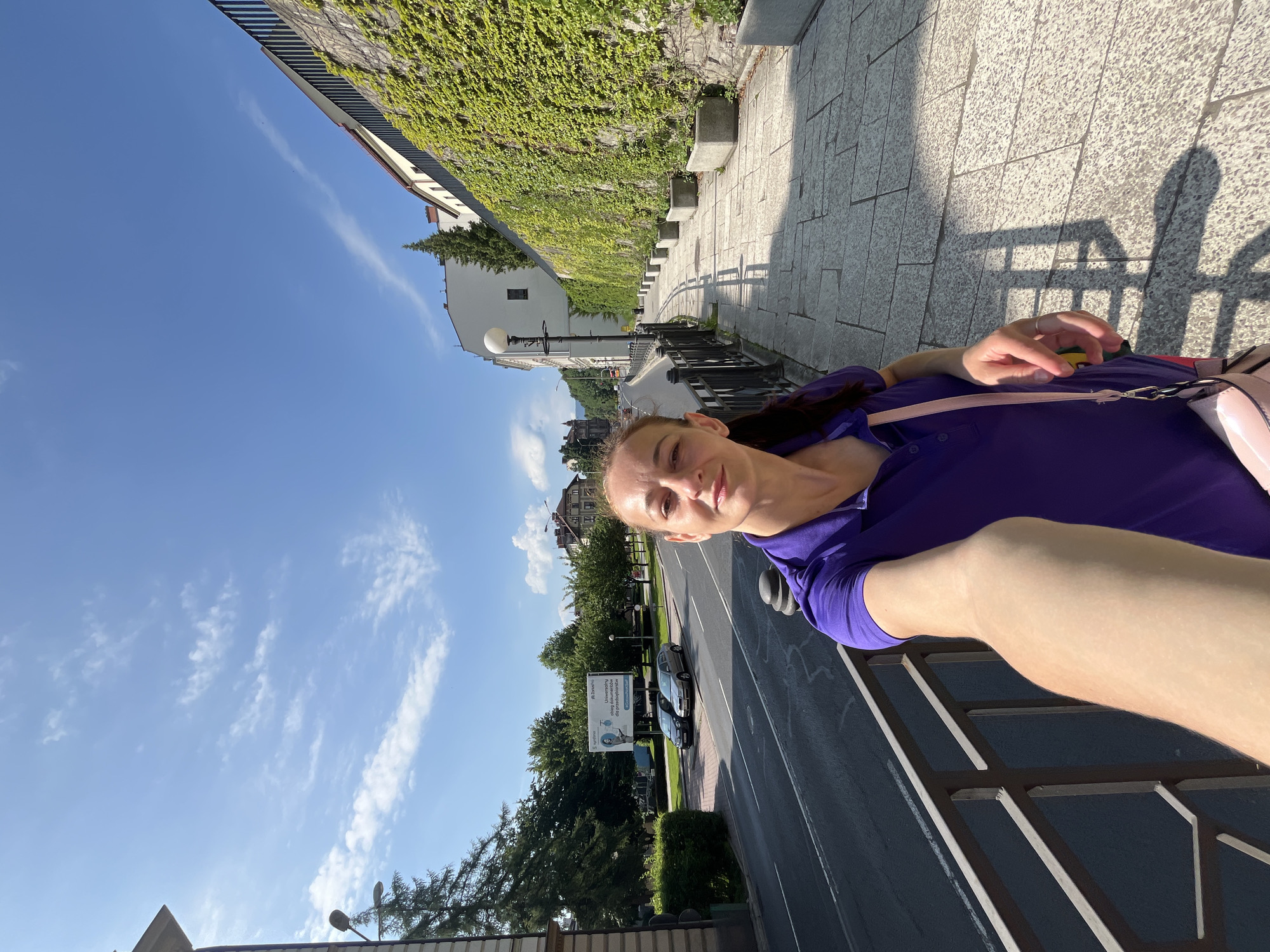 In the city of Unknown, Unknown, on a clear, sunny day, a woman in a purple polo shirt stands on a paved sidewalk, holding her phone to take a selfie. She has her left arm extended, and her face is partially visible. To her right, a stone wall covered in vibrant green ivy runs alongside the sidewalk, punctuated by concrete planters. A black metal railing separates the sidewalk from a street on the left, where two cars are parked near a billboard. The street continues into the distance, lined with trees and buildings, under a bright blue sky with scattered white clouds.