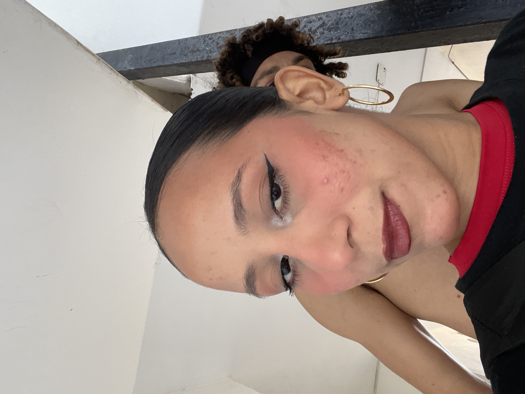 The image is a selfie taken indoors, likely in an urban residential setting in Porlamar, Venezuela. The primary subject is a young woman with dark hair pulled back, facing the camera. She is wearing dark lipstick, eyeliner, and blush. She has acne on her cheeks and a gold hoop earring in her visible ear. Behind her, partially obscured by her shoulder, is another person with curly dark hair and a black headband, peeking from behind a dark metal railing. The background consists of a white wall and what appears to be part of a ceiling or ledge, with some discoloration and dust visible. The overall lighting suggests an indoor environment with ambient light.