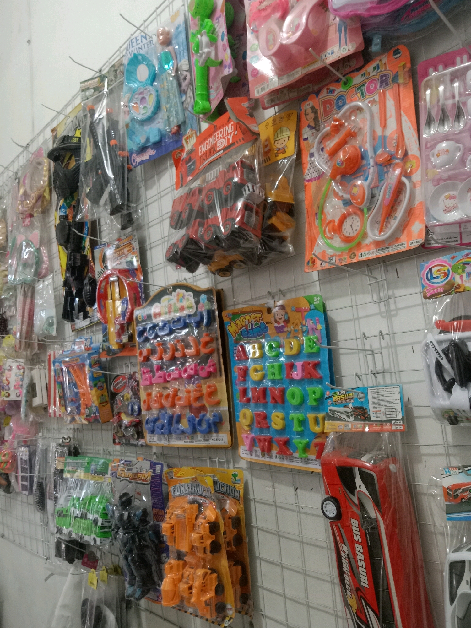 The image shows a wall display of various toys in what appears to be a small shop or market stall in Sugihwaras, Indonesia. The toys are hung on a wire grid. On the top shelf, there are toy makeup kits, dolls, and toy doctor kits. Below those, there are toy guns, toy vehicles, and educational toys like alphabet blocks. The lighting is artificial, and the wall behind the display is white. The overall impression is of a retail environment selling children's toys.