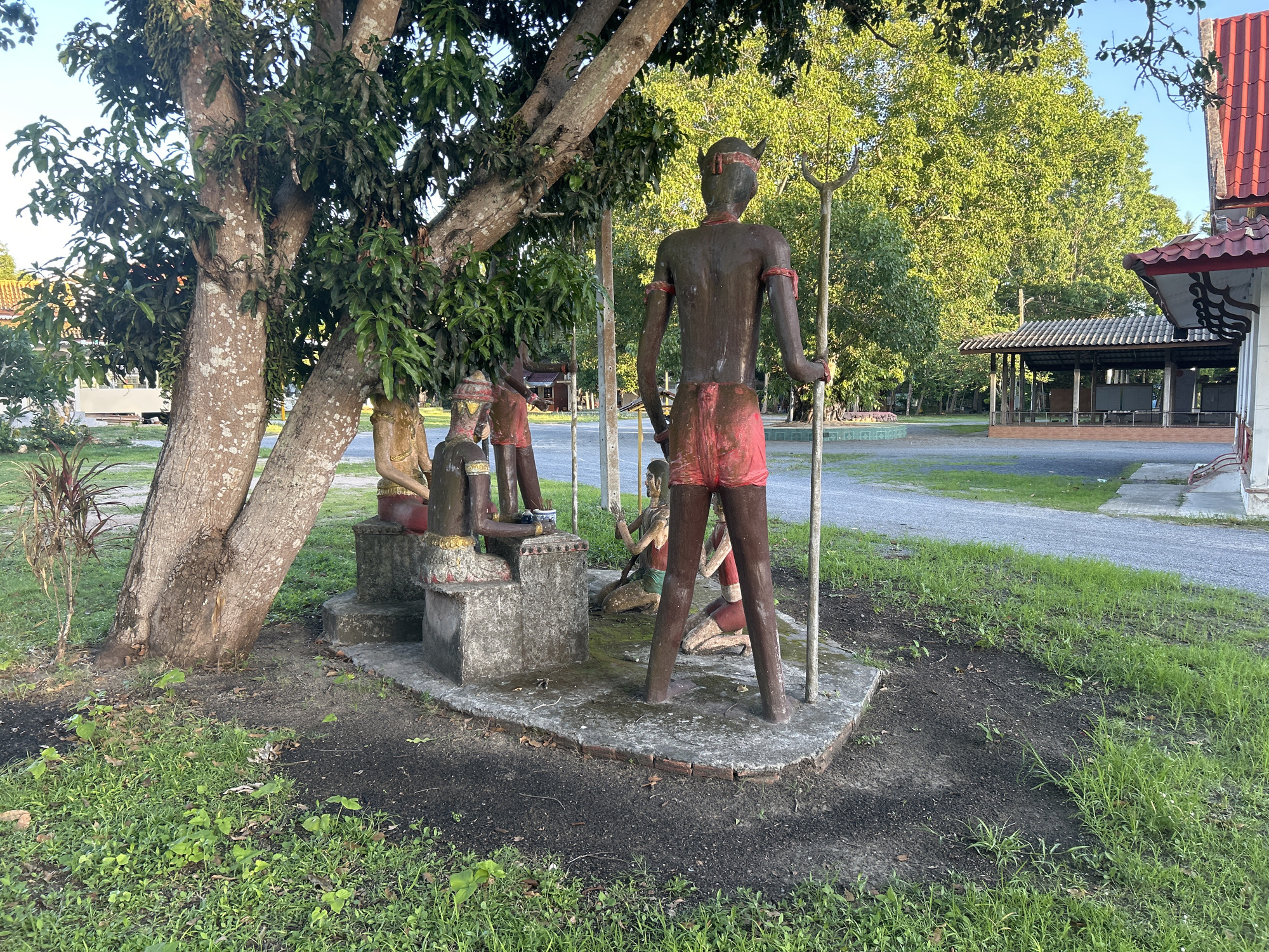 The image depicts a group of statues placed on a concrete pedestal under the shade of a large tree. The statues appear to represent human figures, predominantly with dark skin tones and adorned with red garments, possibly loincloths and headbands. One prominent statue in the foreground stands facing away from the camera, holding a pole-like object with a trident-like end. To its left, other statues are arranged, some in seated or kneeling positions. The setting is outdoors, with lush green grass and foliage surrounding the statues. In the background, a road is visible, along with buildings featuring red tiled roofs and a covered structure. The overall atmosphere suggests a cultural or religious site located in ตำบลเกาะพะงัน, Thailand. The lighting indicates it might be late afternoon or early morning, with sunlight filtering through the leaves of the tree and illuminating the scene.