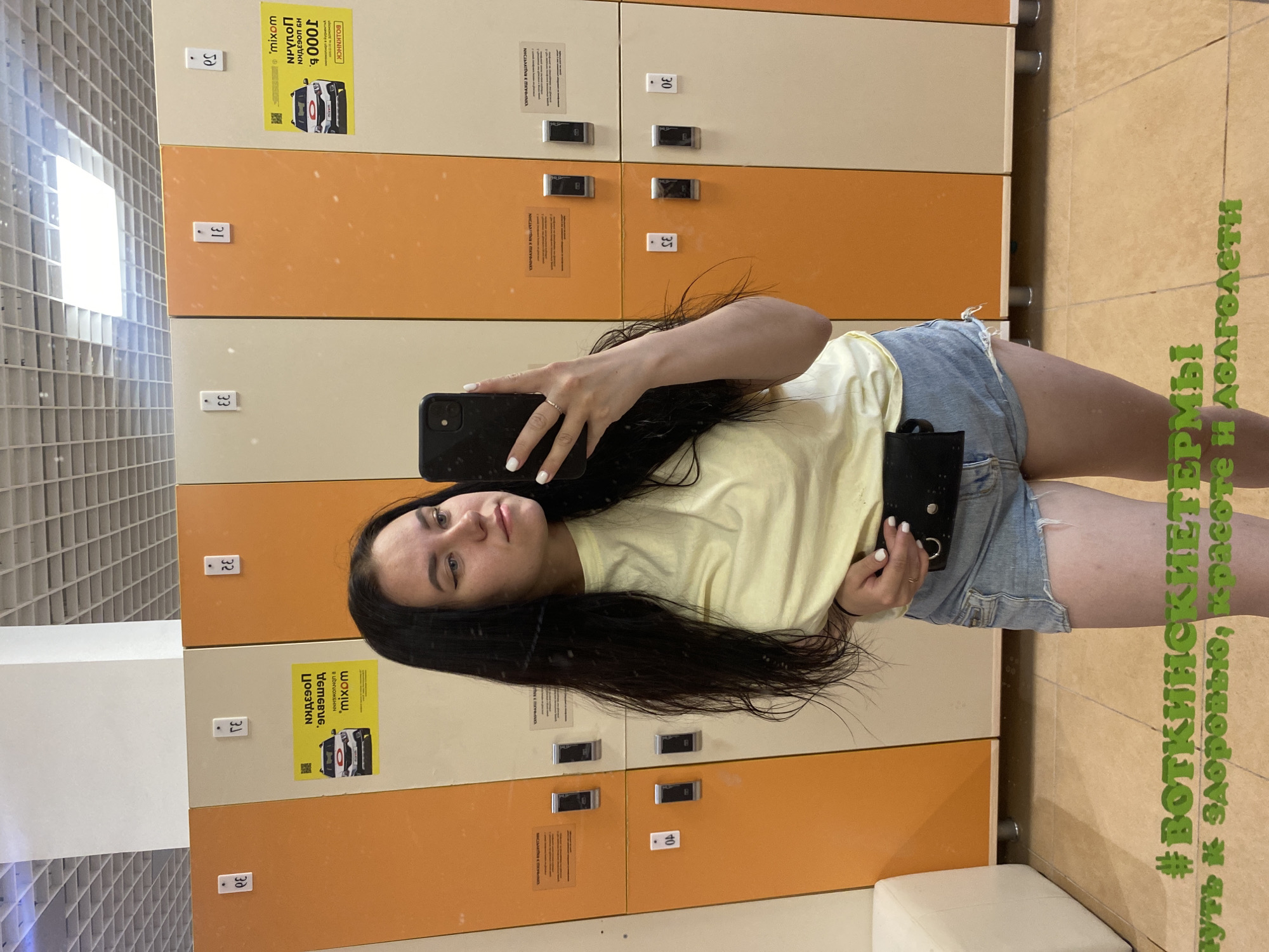The image shows a woman taking a selfie in a locker room at the Votkinskiye Termy in Votkinsk, Russia. She is holding a phone and a small black object, possibly a wallet or cardholder. The lockers are a mix of orange and beige, numbered from 30 to 50. Advertising is visible on the lockers. The walls and floor are tiled. The environment appears to be a public changing area.