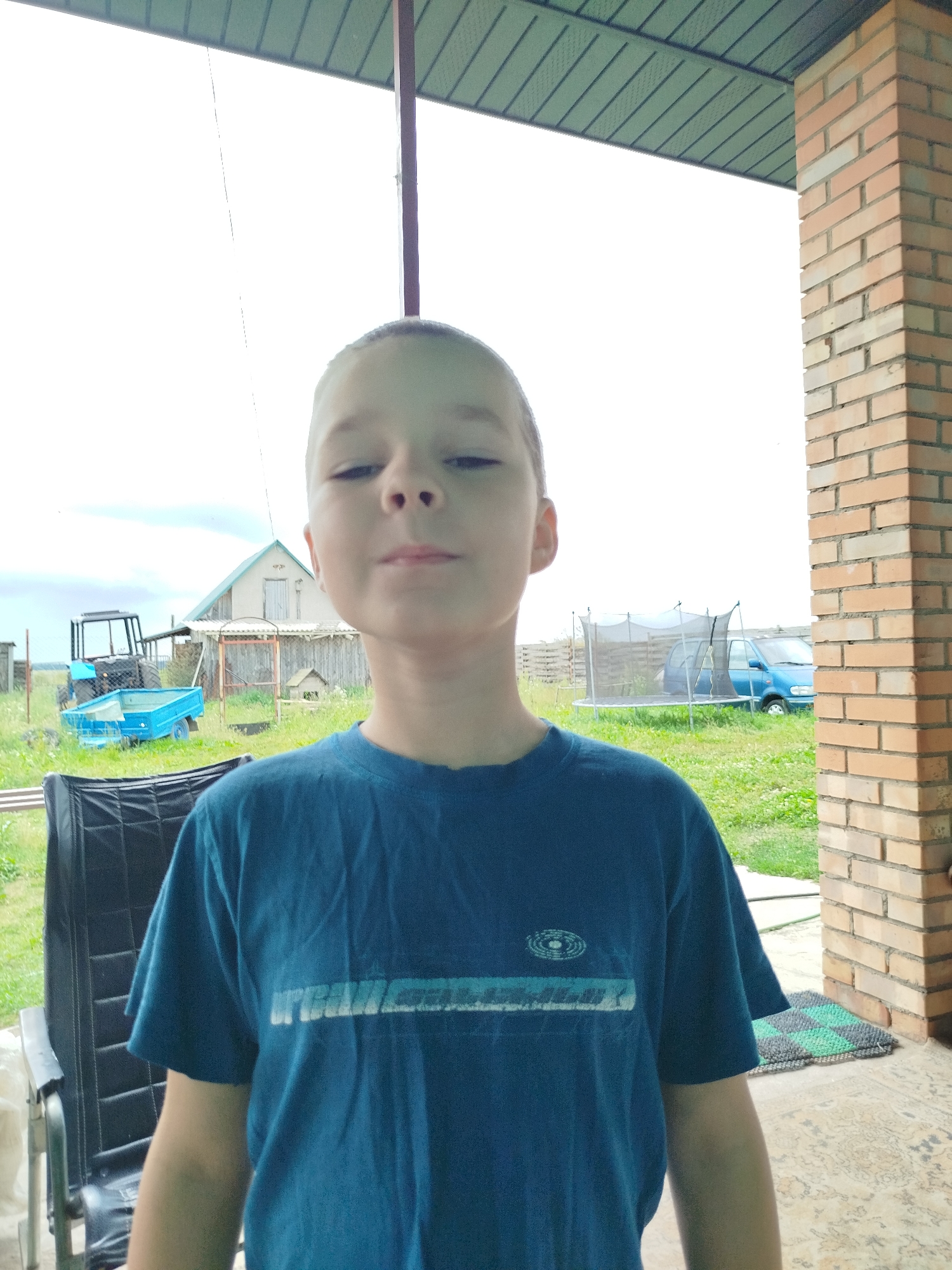 A young boy in a blue t-shirt is standing outside a rural building in Крывасельскі сельскі Савет, Belarus. He is facing the camera, looking directly at it. The background shows a grassy yard with a blue tractor, a blue trailer, a house, a trampoline, and a blue car. A brick column and part of a roof are visible to the right of the boy. The overall scene suggests a casual, outdoor setting in a village.