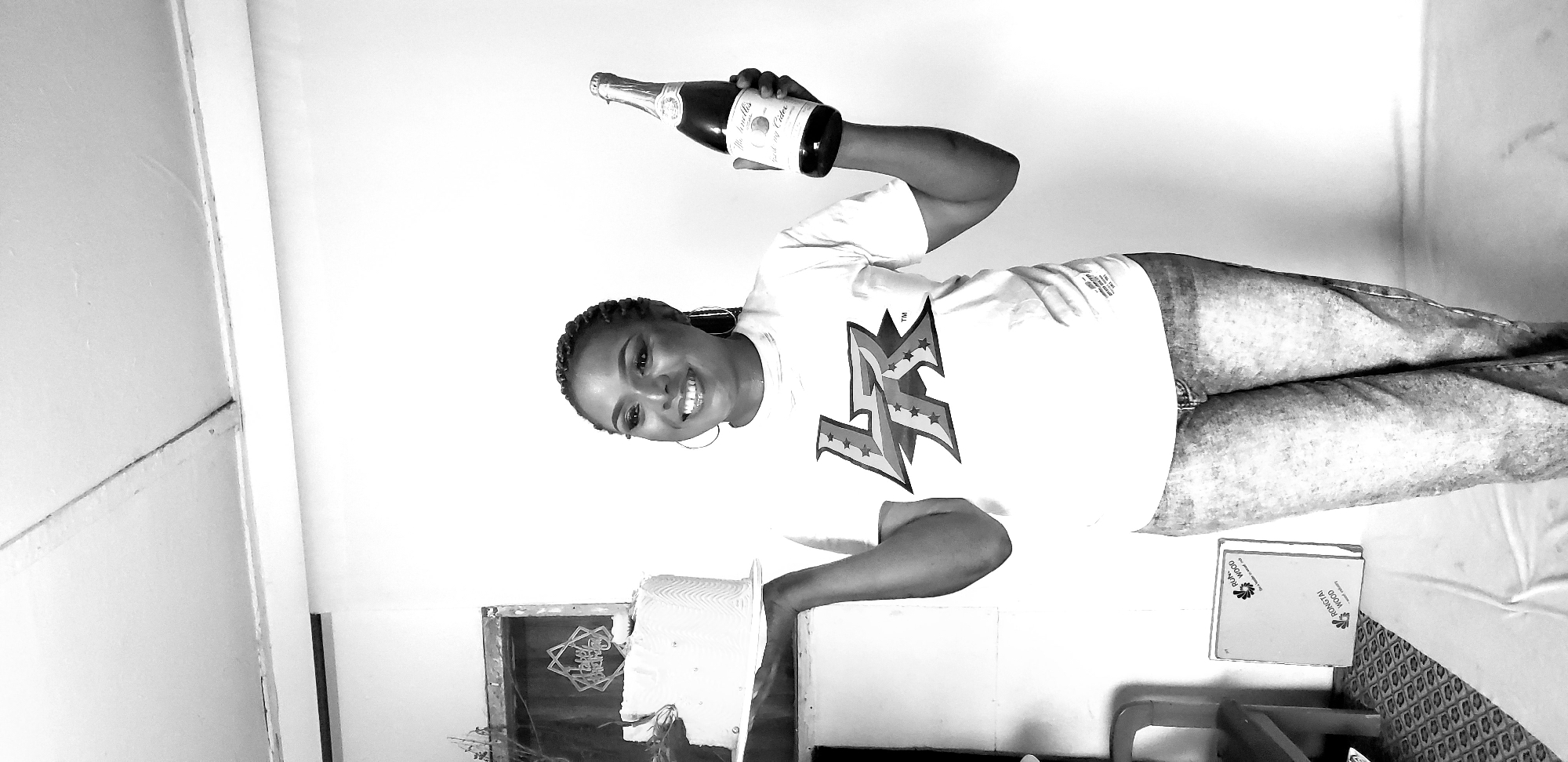 A full-body, black and white, medium shot captures a young Black woman standing against a plain white background, seemingly in a studio environment or well-lit room in Sabon Gari, Nassarawa, Nigeria. She is holding a white cake with a gold topper and a bottle of sparkling cider. The woman has dark, braided hair pulled back, large hoop earrings, and is wearing a white t-shirt with a graphic logo and distressed denim jeans. Her makeup is dramatic, with a bold lip and winged eyeliner. The background is a bright white wall, with a small portion of a patterned floor visible at the bottom. To the left, a chair and a framed object are slightly visible, suggesting a staged setting. The overall impression is one of celebration and a photoshoot.