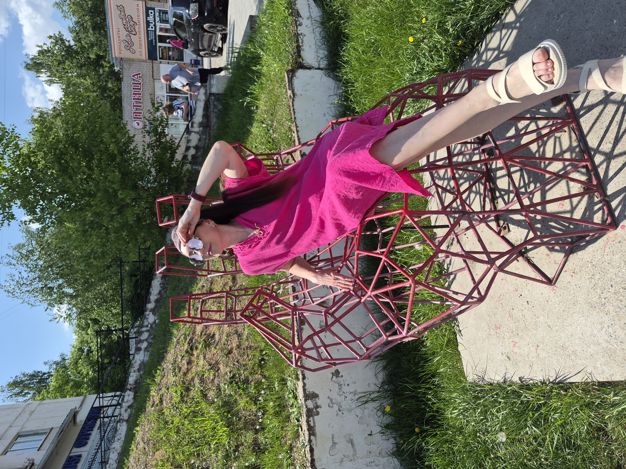 A woman is reclining in a red, geometric metal sculpture, possibly a chair, in a public outdoor space. She is wearing a bright pink dress and white sandals, sunglasses, and a headband, and has long dark hair. Her head rests on her hand, and she appears relaxed. The sculpture sits on a concrete slab surrounded by grass. In the background, there is a building with signs in a Cyrillic script, trees, and a few people walking nearby. A car is also visible in the background. The location seems to be a city in Russia, based on the Cyrillic script on the building. The environment is sunny.